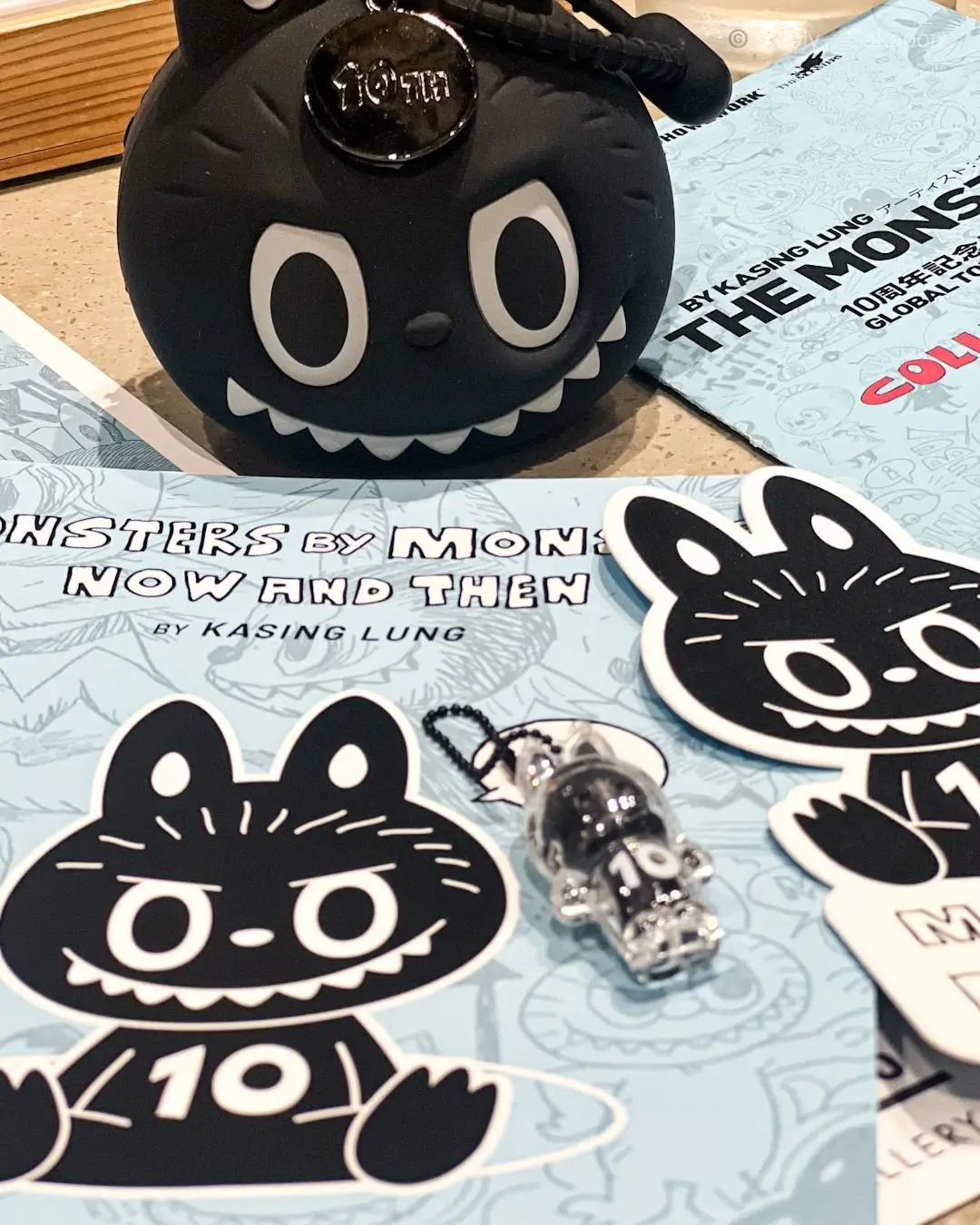 黒い猫の顔のデザインが施されたぬいぐるみとステッカー、クリアのキーホルダーが青い背景の上に並んでいる
