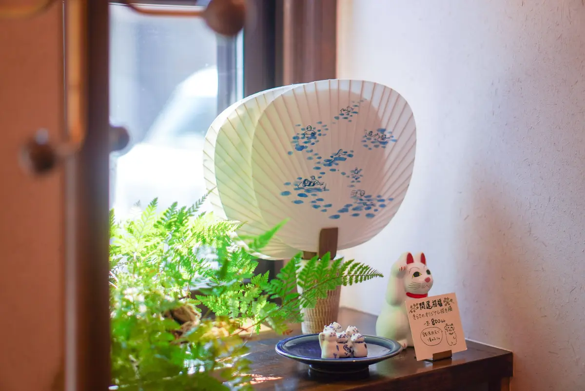 窓辺に置かれた、扇子、苔むした盆栽、招き猫の装飾。扇子は白地に青色の魚のデザイン。盆栽は緑豊かな苔と植物で覆われ、招き猫は台座に乗って立っている。