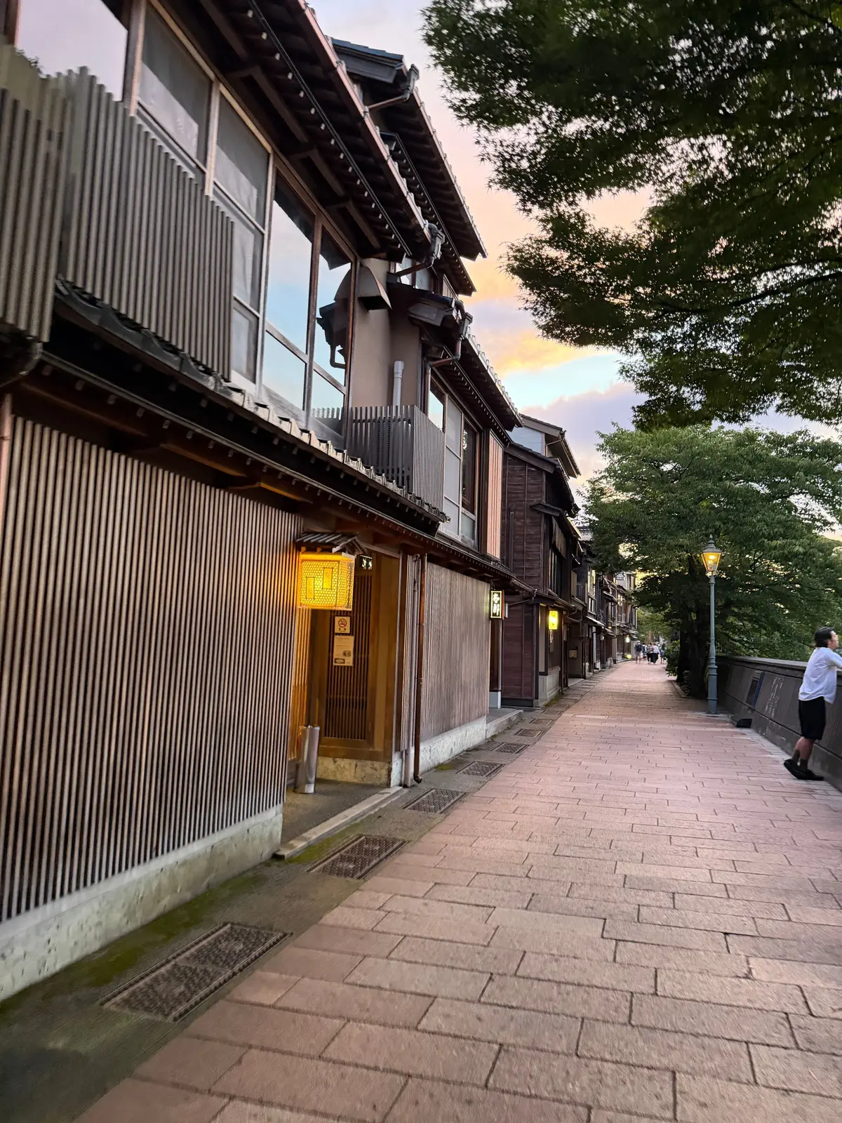 夕暮れ時の日本の街並み。両側に木造の伝統的な家並みが並び、石畳の道が奥へと続いている。緑豊かな木々が背景にあり、空はオレンジ色に染まっている。