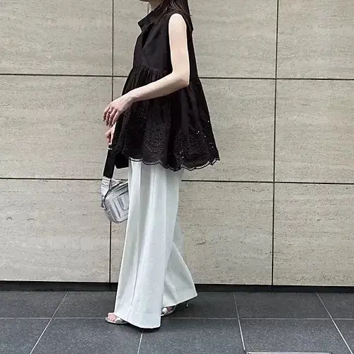 黒いレースのトップスと白いワイドパンツを着用した女性。右手にバッグを持ち、横断歩道を歩いている。背景はベージュ色のタイル壁。