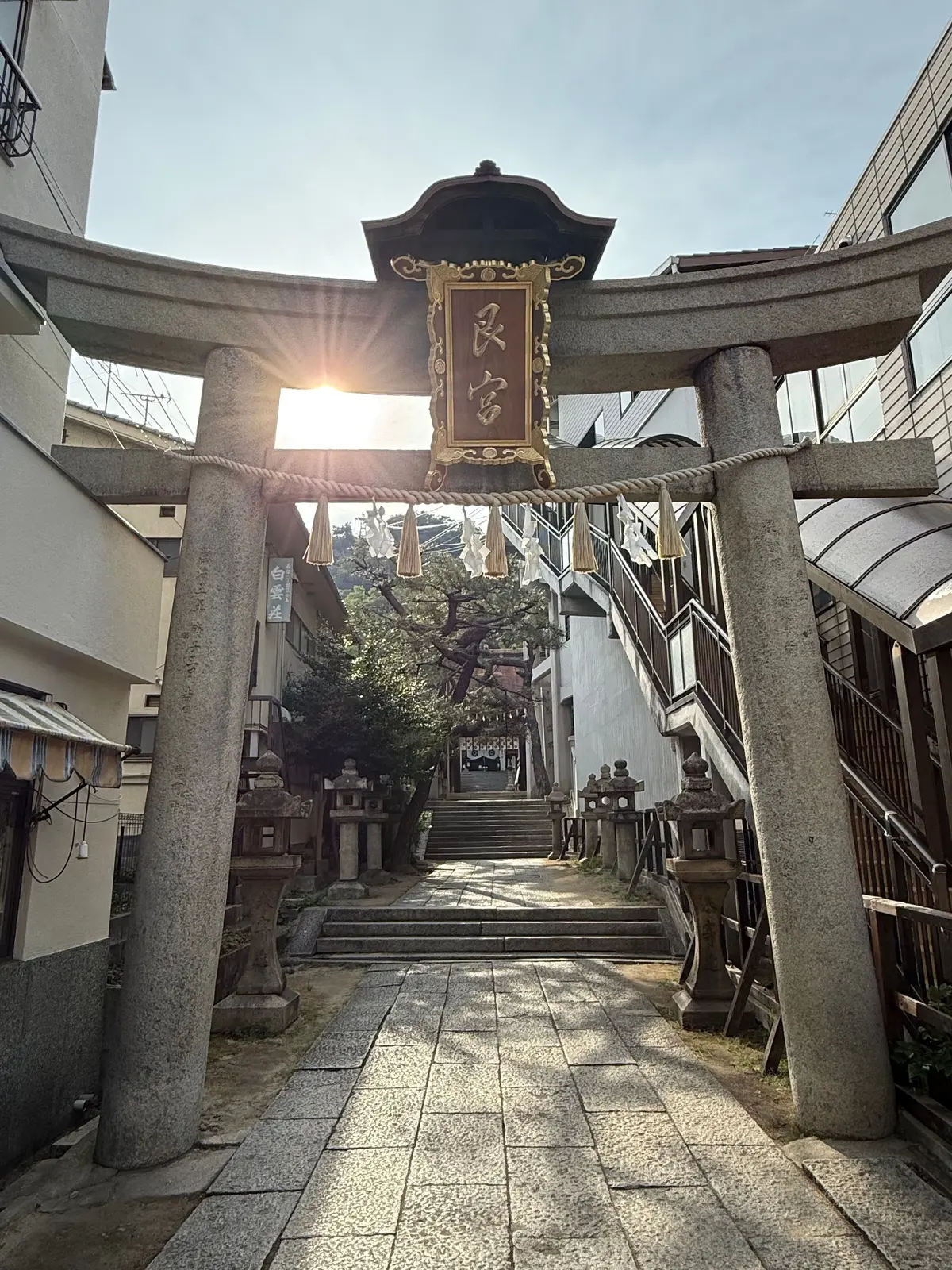 石造りの鳥居がアーチ状に配置され、その上には赤い額に漢字が書かれている。鳥居の先には階段が続き、陽光が差し込んでいる。周囲には建物が並ぶ。広島、尾道で、尾道観光。尾道三部作の舞台巡り。まずは、時をかける少女の舞台の艮神社。