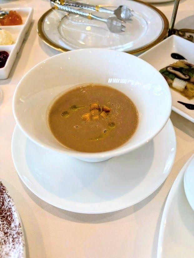 白い器に入った、茶色のスープ。表面には、細かく刻んだナッツが散らされている。白い皿の上に置かれ、上品なテーブルコーディネートが施されている。