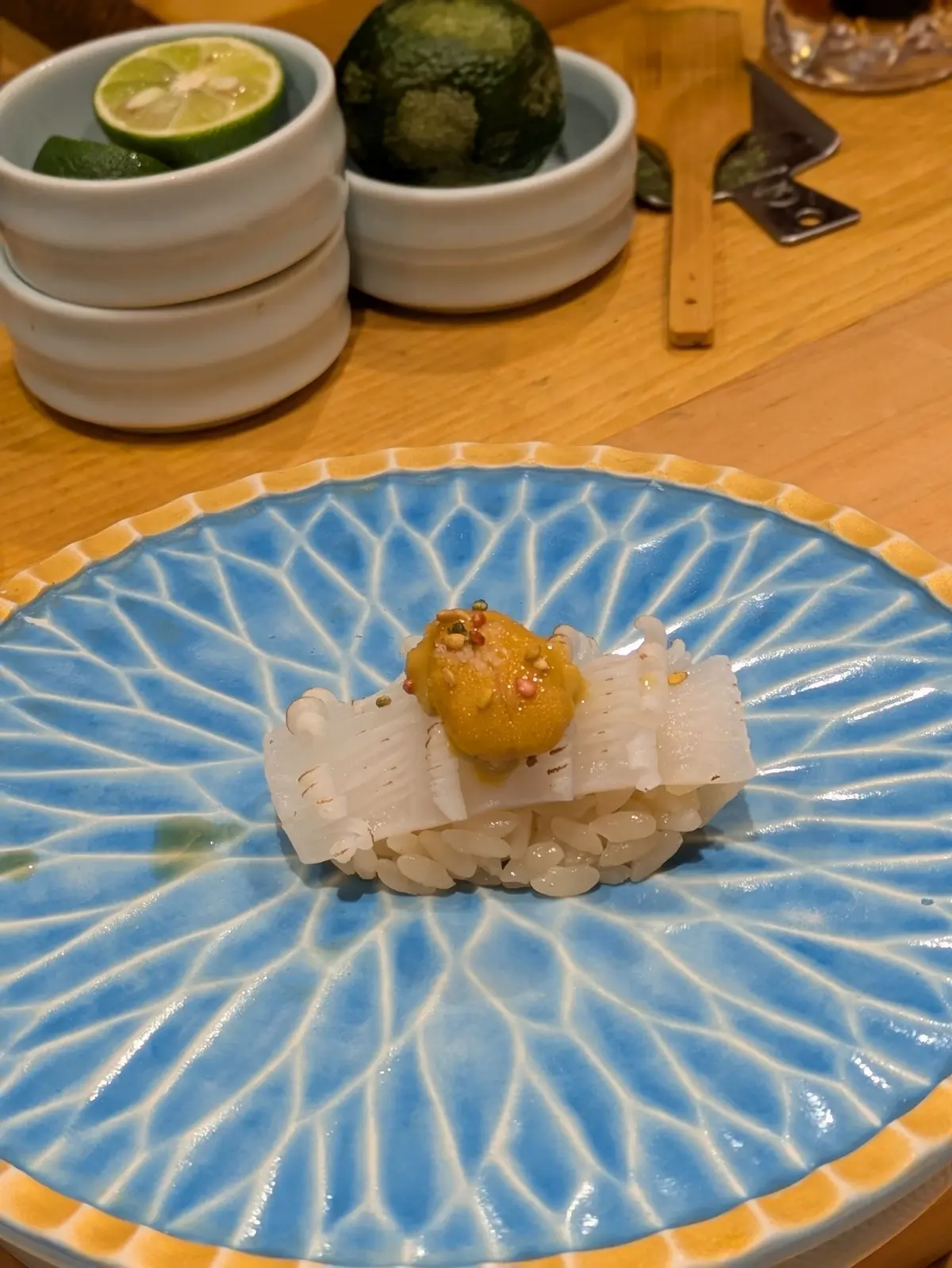 青い波模様の器に、薄い白身の魚の切り身と黄色いソースがのった料理。木製のテーブルに、ライムとアボカドの小鉢が並んでいる。