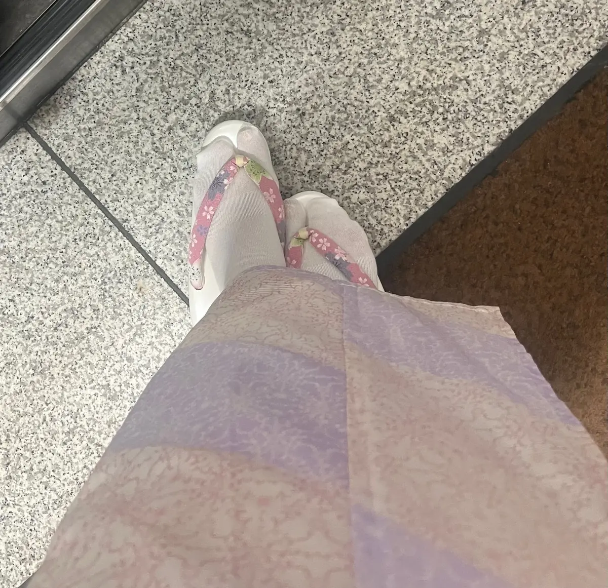 白いゲタと薄紫色の透け感のあるスカートが写っている。ゲタの履き口には花柄の装飾がある。背景はグレーのタイルの床。