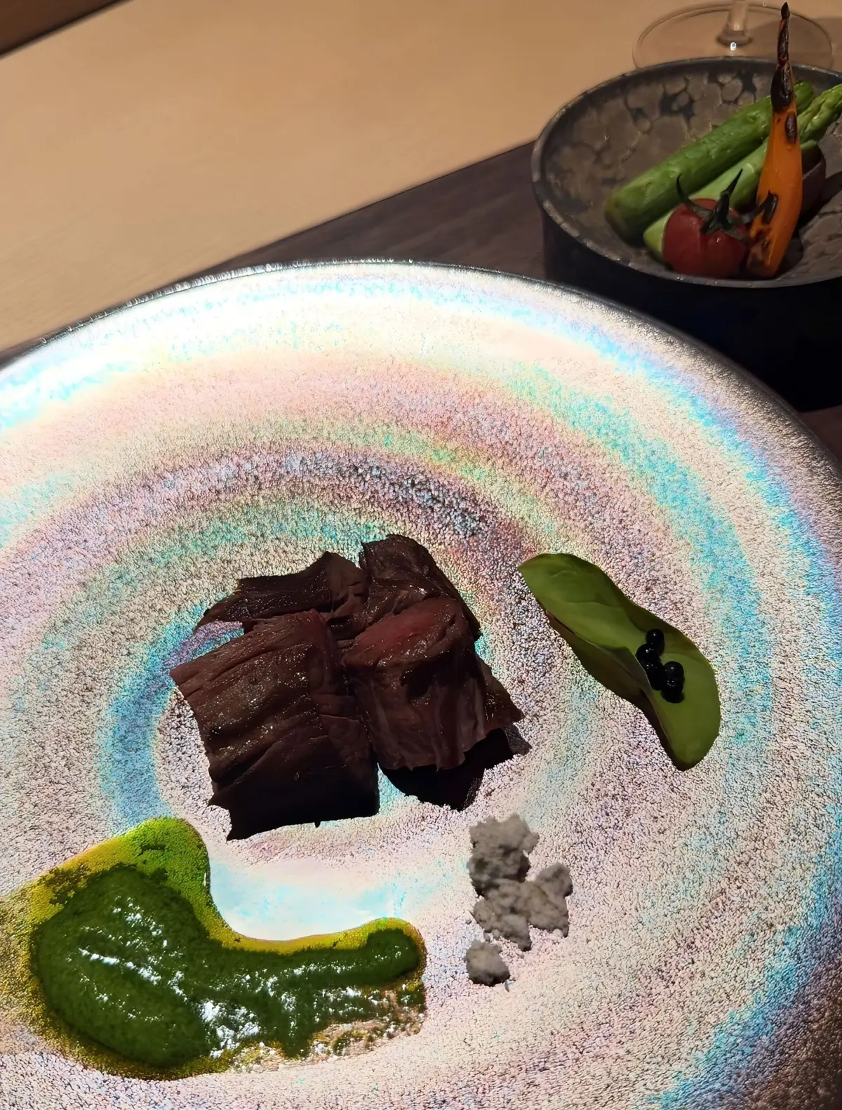 高級レストランの皿に盛り付けられた料理。中央には赤褐色の肉が数切れ、左下には緑色のソース、右下には白い粉状のものが置かれている。皿は虹色の模様が施され、右には野菜の小鉢が添えられている。