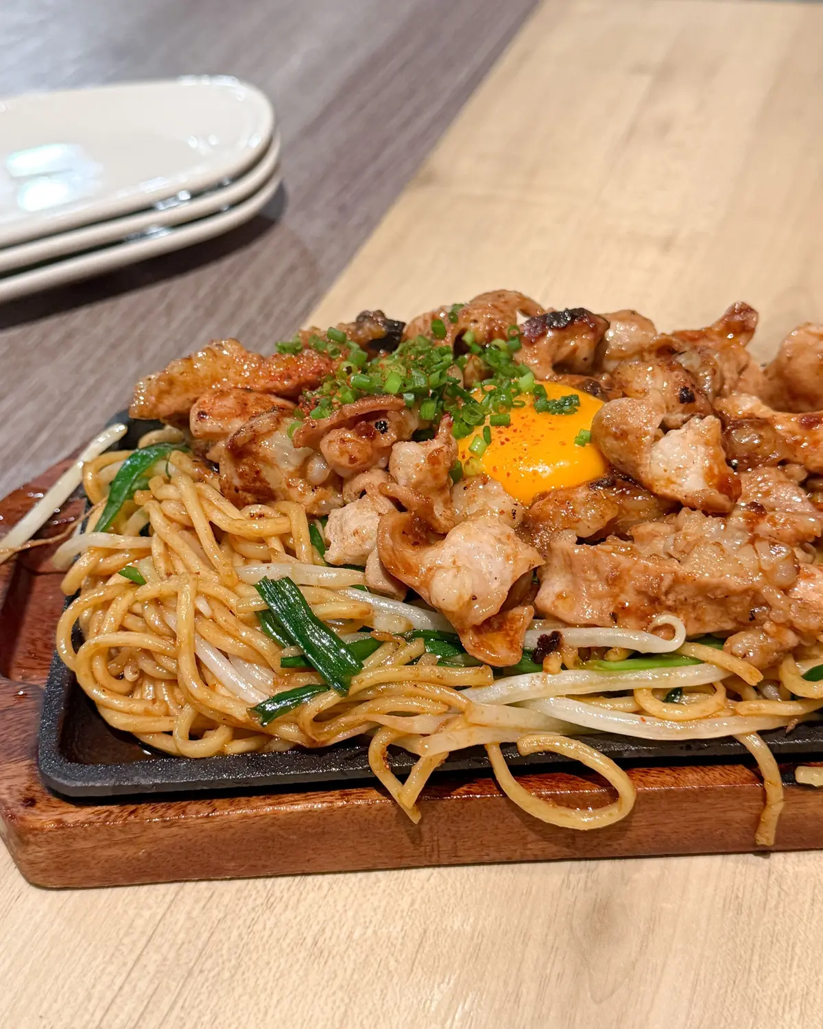 木製のプレートに盛り付けられた、豚肉と卵黄を乗せた焼きそば。麺は黄色く、豚肉はカリカリに焼かれており、ネギと豆芽が添えられている。