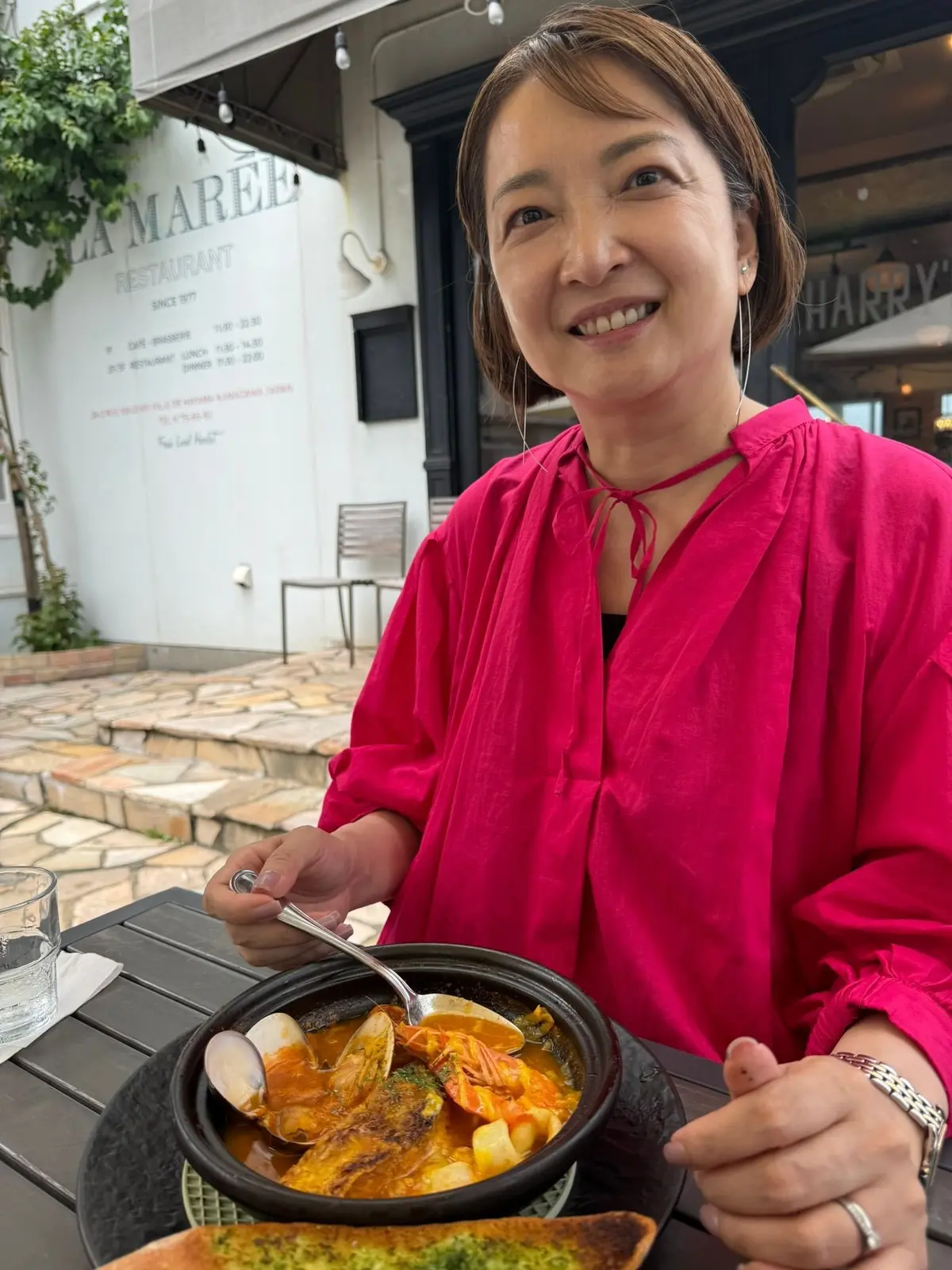 ピンクのブラウスを着た女性が、屋外のレストランで黒い鍋に入った料理をスプーンで食べている。背景には「La Marel Restaurant」の看板が見える。