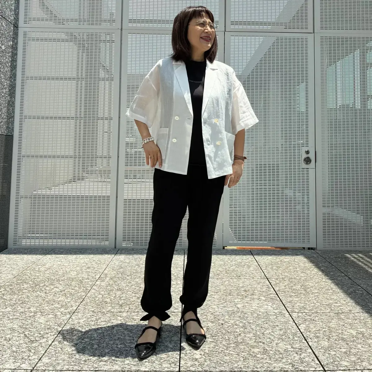 黒いトップスに黒いパンツ、白いリネンジャケットを羽織った女性。眼鏡をかけ、腕には腕時計とブレスレット。黒いストラップシューズを履き、背景には白い金属製の壁がある。