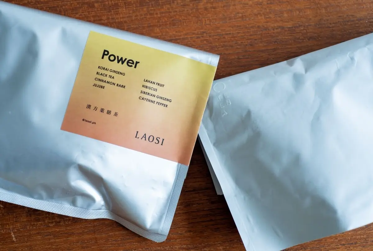 木製のテーブルに置かれた、LAOSI社の「Power」という名のハーブティーの袋。茶色と白のストライプ模様で、成分リストが記載されている。隣には、同じブランドの白い袋が置かれている。