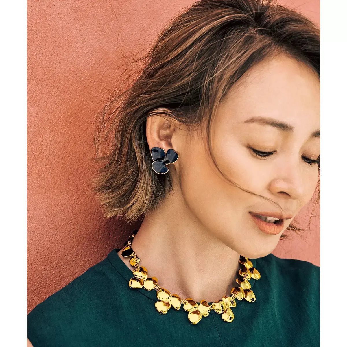 ダークグリーンの服を着た女性で、左耳に青い花びら型のイヤリング、右耳に金色の花びら型のイヤリングを着用。首元には金色の花びら型のネックレスを身につけている。