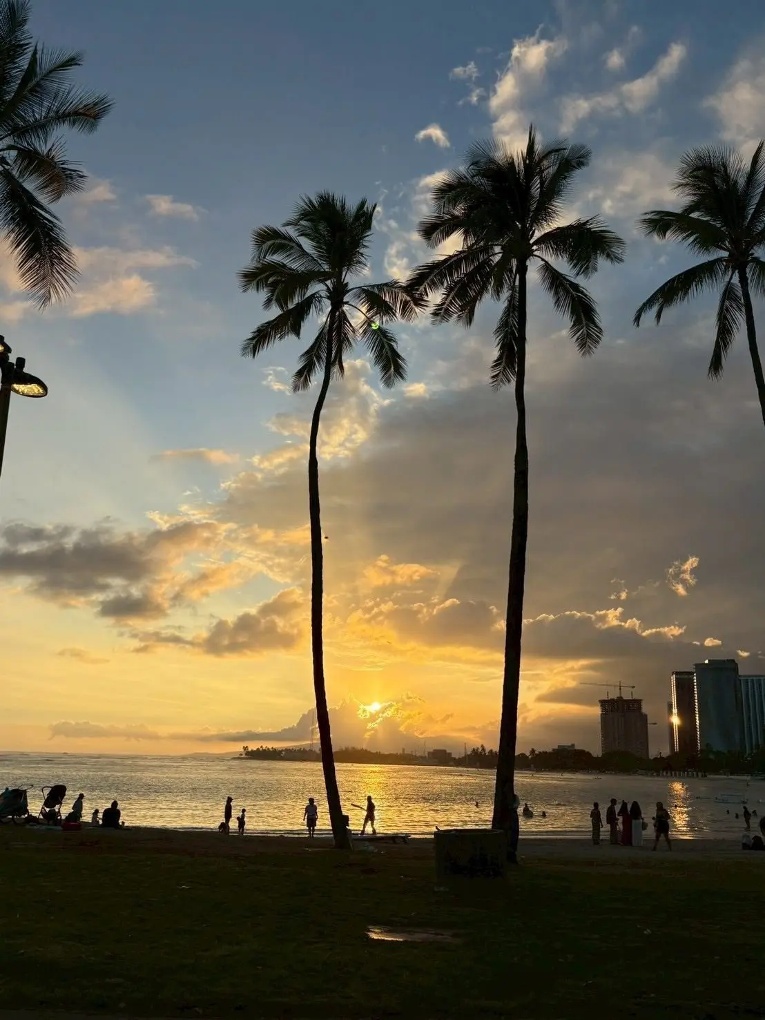 夕日が海に沈むビーチの風景。ヤシの木が並び、人々が海辺で散歩する。遠くには高層ビルが映る。