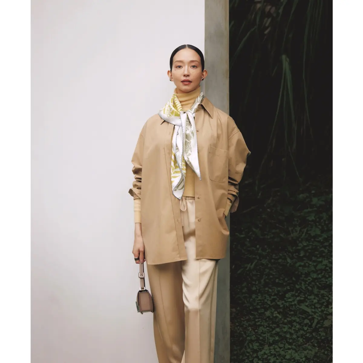 ベージュのタートルネックニットの上にシャツをはおり、センタープレスパンツをあわせたワントーンコーディネートに、白×イエローのプリントスカーフをアクセントとして首元に巻いたスタイリング　モデル・松島花