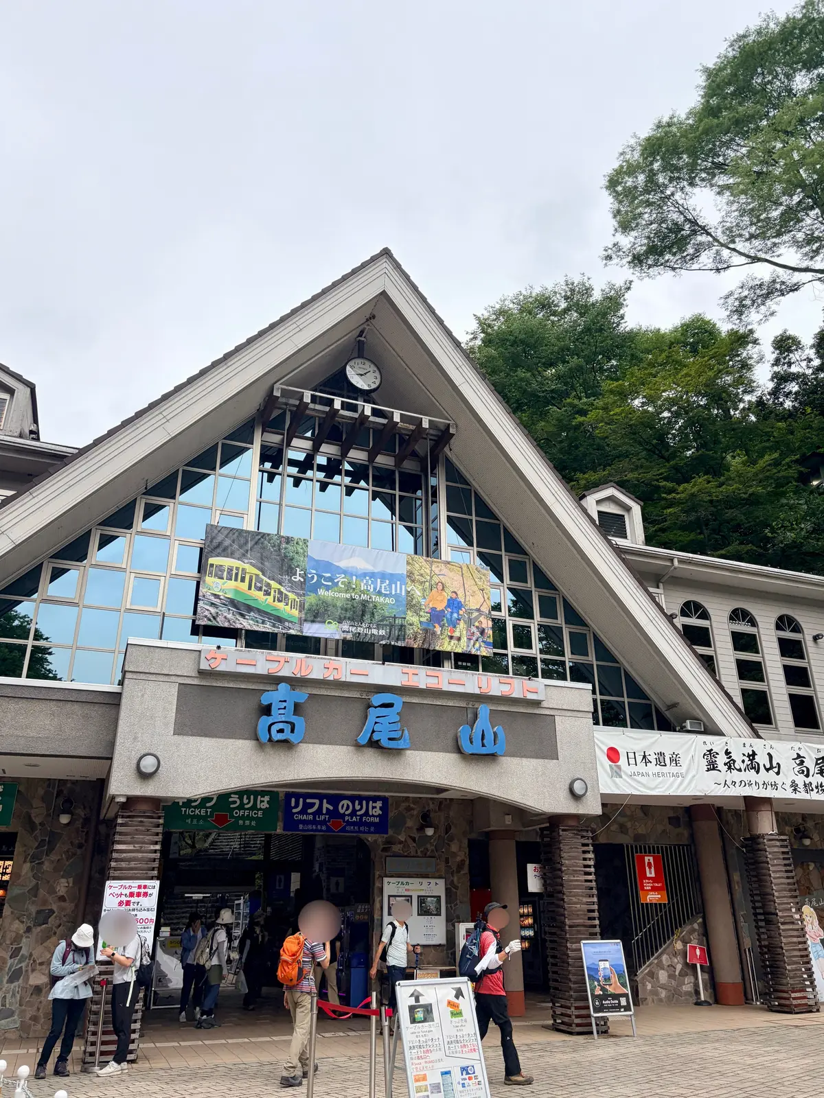 高尾山ケーブルカーの乗り場、ガラス張りの建物と石造りの柱、看板や案内板が設置された広場