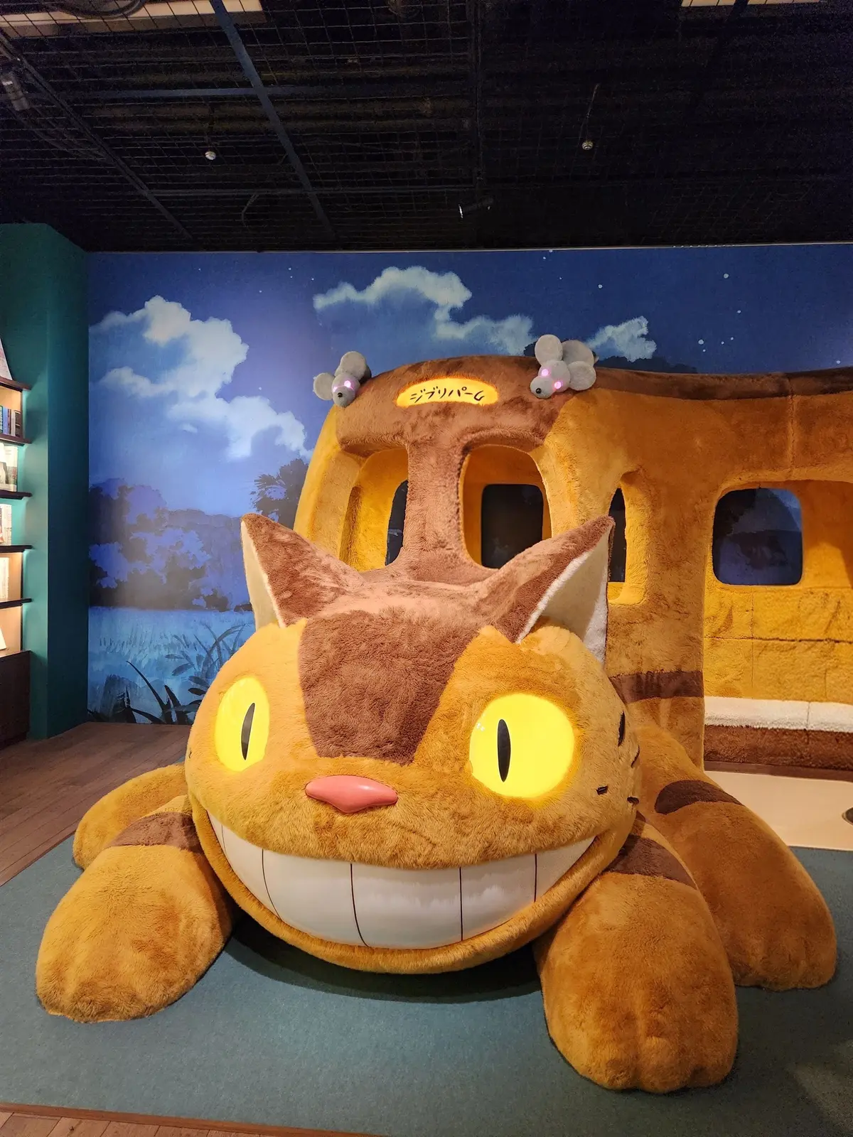 ジブリ作品「猫の恩返し」に登場する、猫のバスの巨大なぬいぐるみ。黄褐色で、大きな黄色い目と口、耳が特徴的。背景には青空と風景が描かれている。