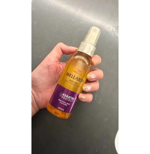 女性の手で保持された、ウェルアトン（Wellaton）のケラチンプロテクト リッチオイルのボトル。ゴールドと紫色のラベルが特徴で、100mlの容量。