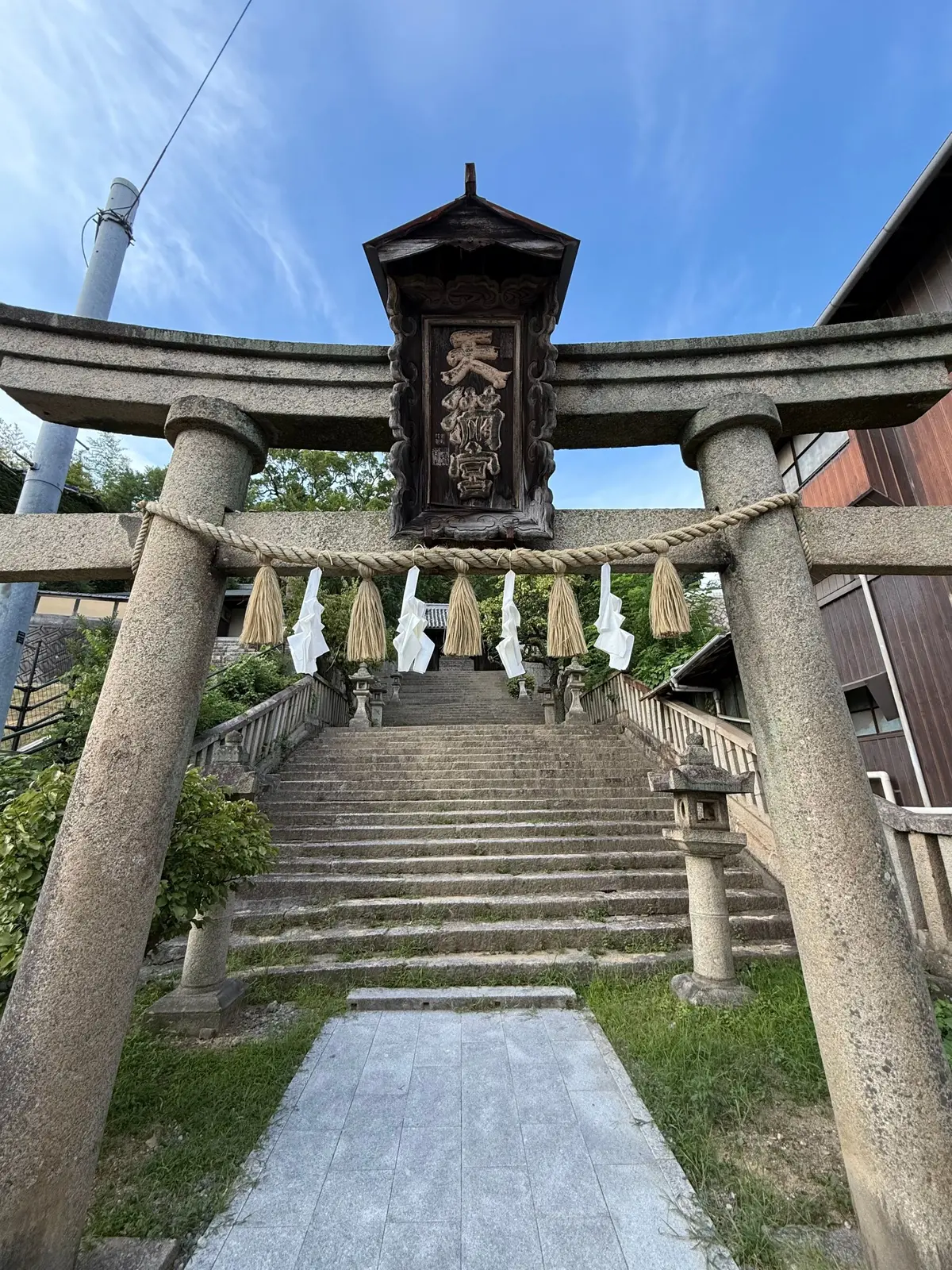 鳥居をくぐり、石段を上がった先にある神社。鳥居は木造で、屋根があり、中央に漢字で書かれた額が飾られている。石段には白い提灯が並び、手すりが設置されている。背景には青空と緑の木々が見える。尾道、御袖天満宮、尾道巡り、尾道観光