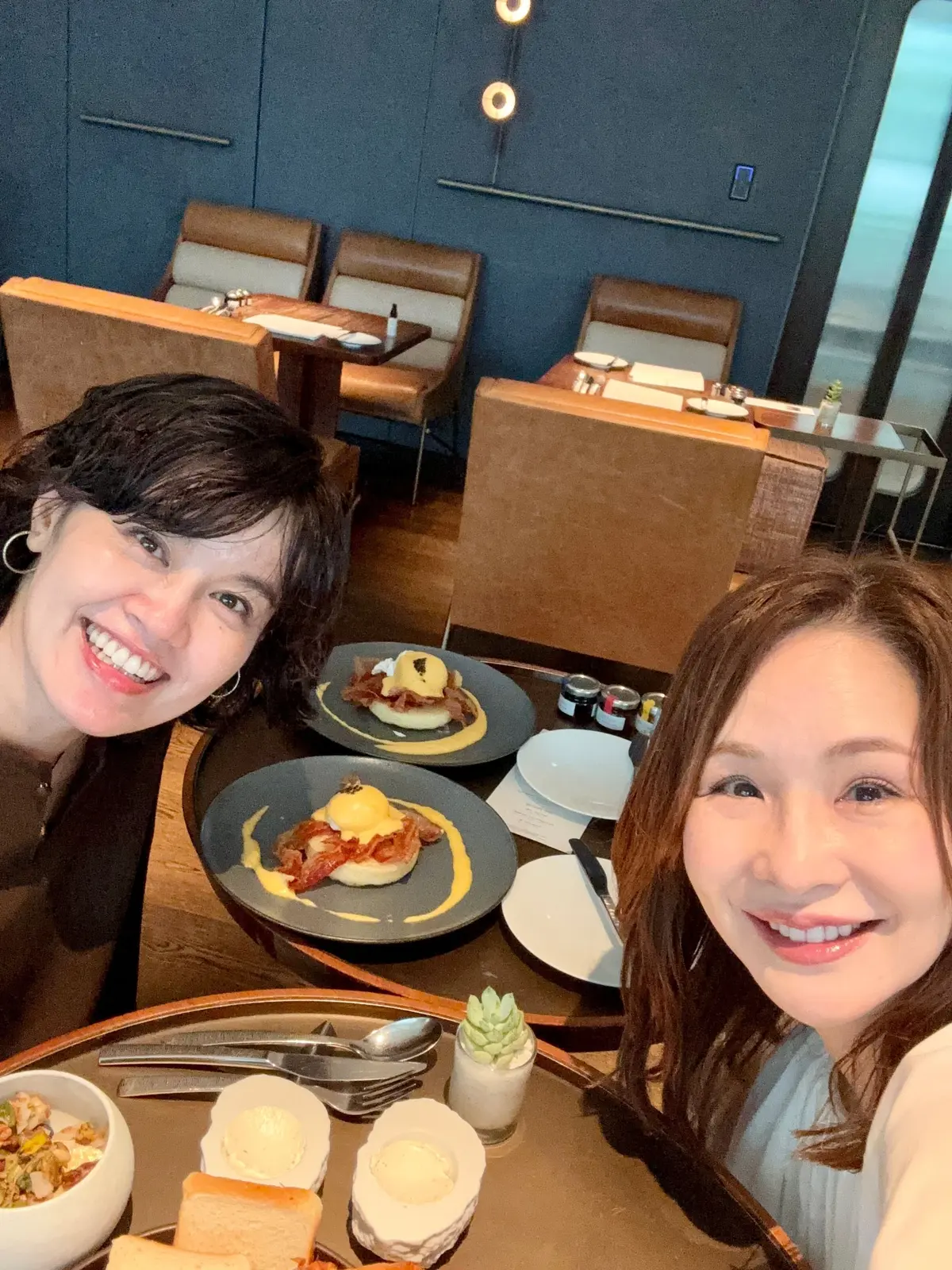 レストランで食事を楽しむ2人の女性。テーブルには、ポテト、パン、チーズ、そしてベーコンとパンケーキの料理が並んでいる。背景には、革張りのソファ席があるモダンな内装が見える。