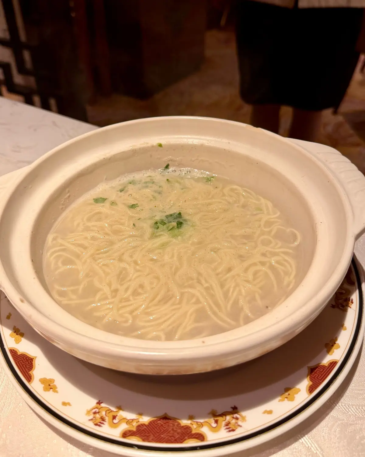 白いボウルに細麺と緑の野菜が乗った淡いスープが盛り付けられ、花模様のプレートに置かれている