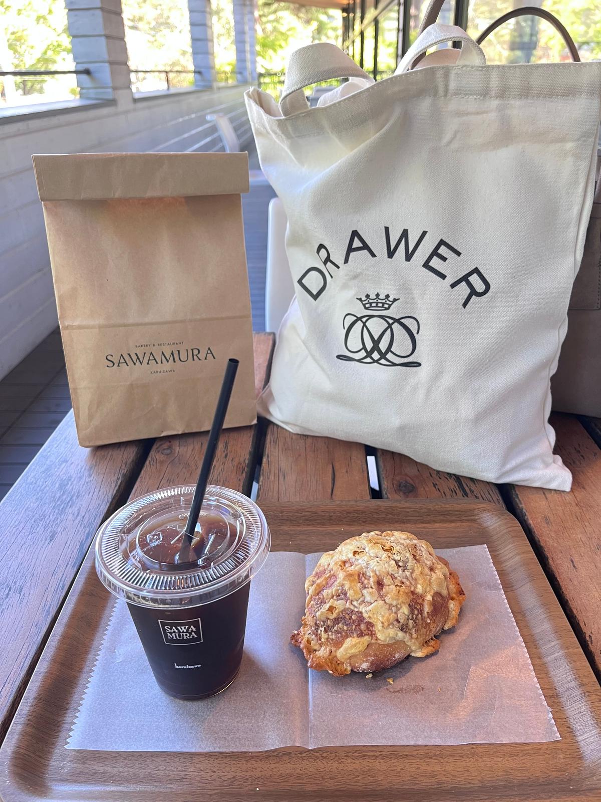 テーブルに置かれた、サワムラ（SAWAMURA）の紙袋、トートバッグ、アイスコーヒー、クリームパン。トートバッグには「Drawer」のロゴがプリントされている。
