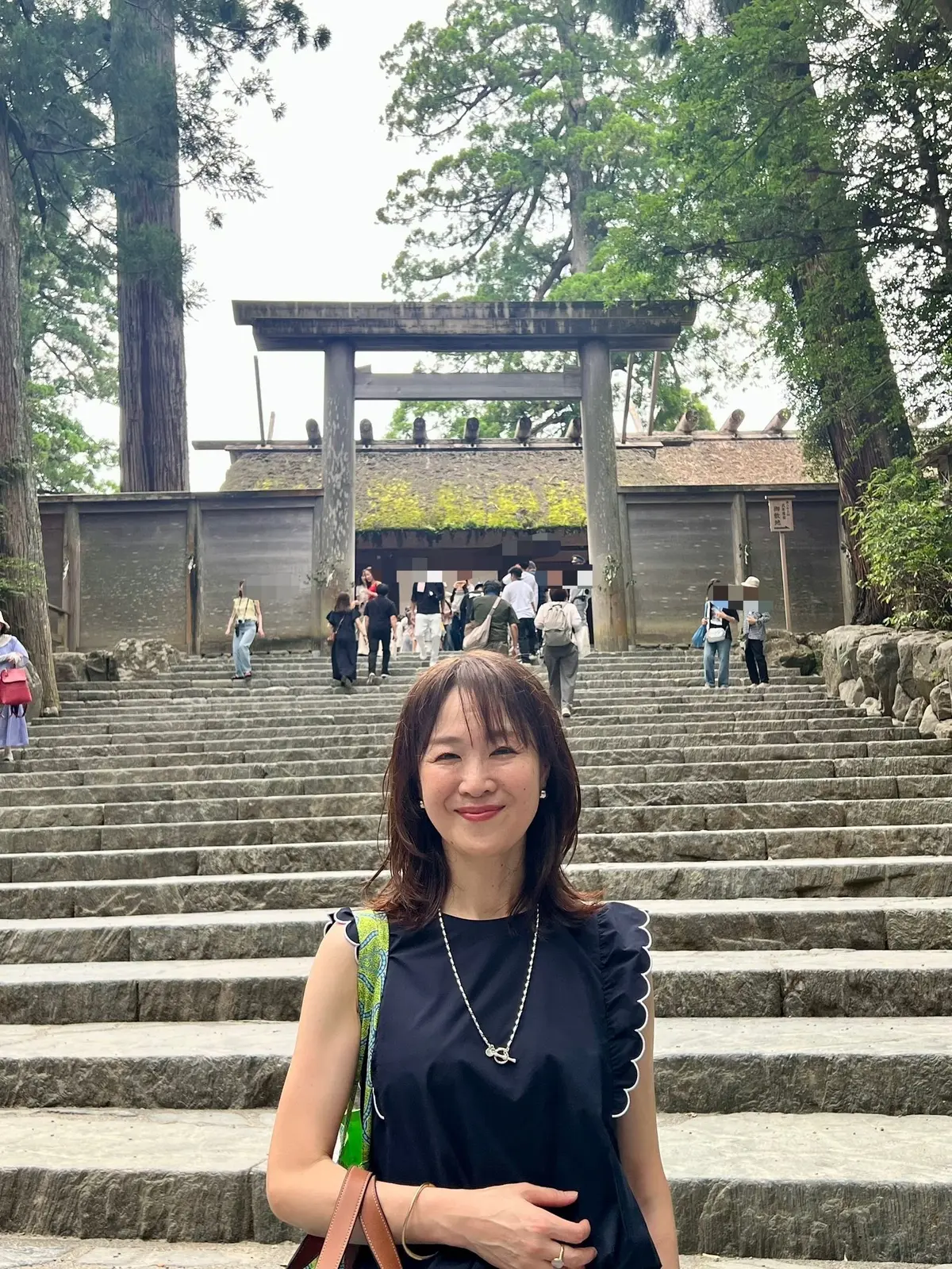 石段と鳥居が続く神社の前に立つ女性。黒いトップスにカラフルなスカーフを着用し、ネックレスを身につけている。背景には緑豊かな木々が見える。