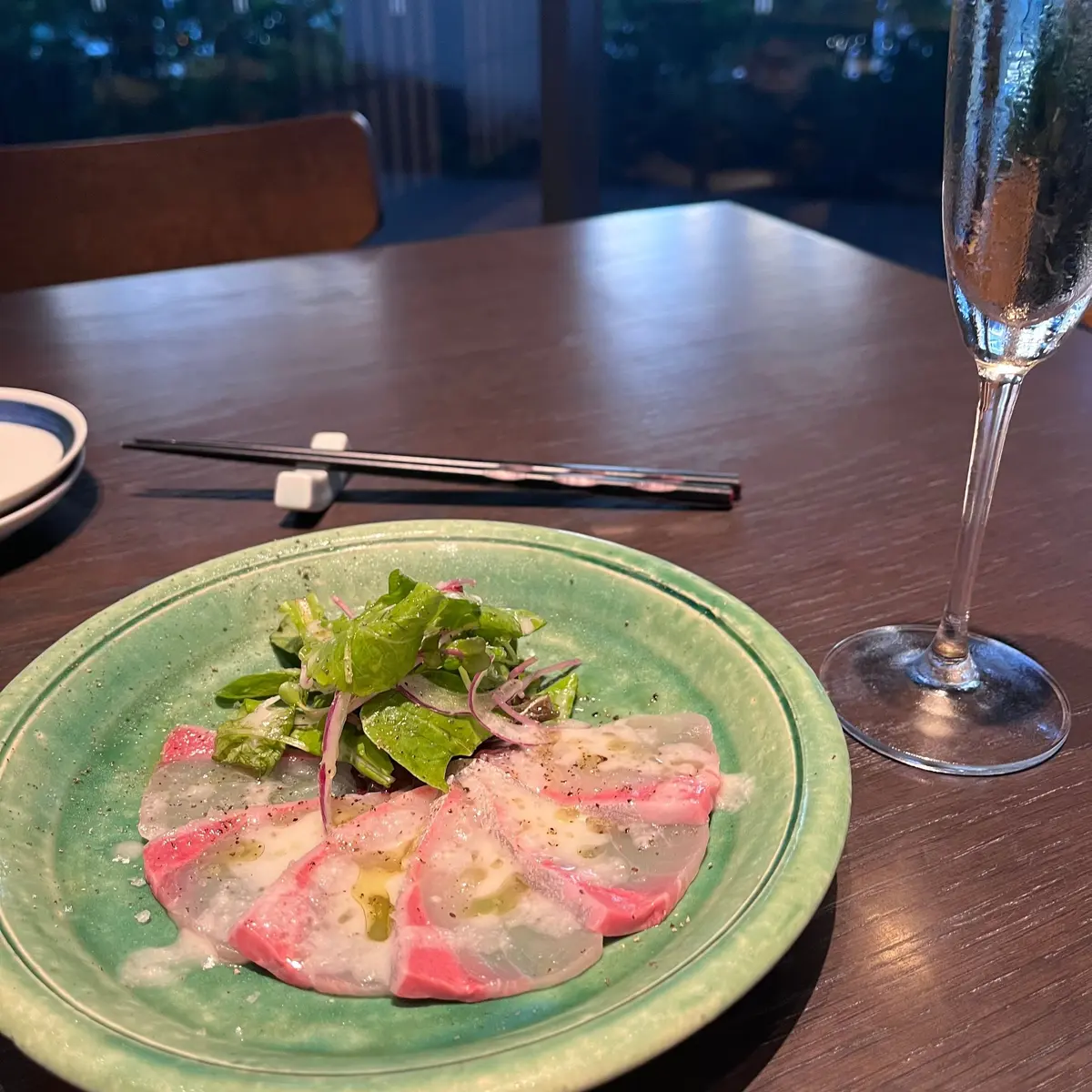 緑色の皿に盛り付けられた、薄切りにしたピンク色の魚介類（刺身のようなもの）。魚介類の上には白いソースと、赤と緑の野菜が添えられている。右隣にはシャンパングラス、手前には箸が置かれている。