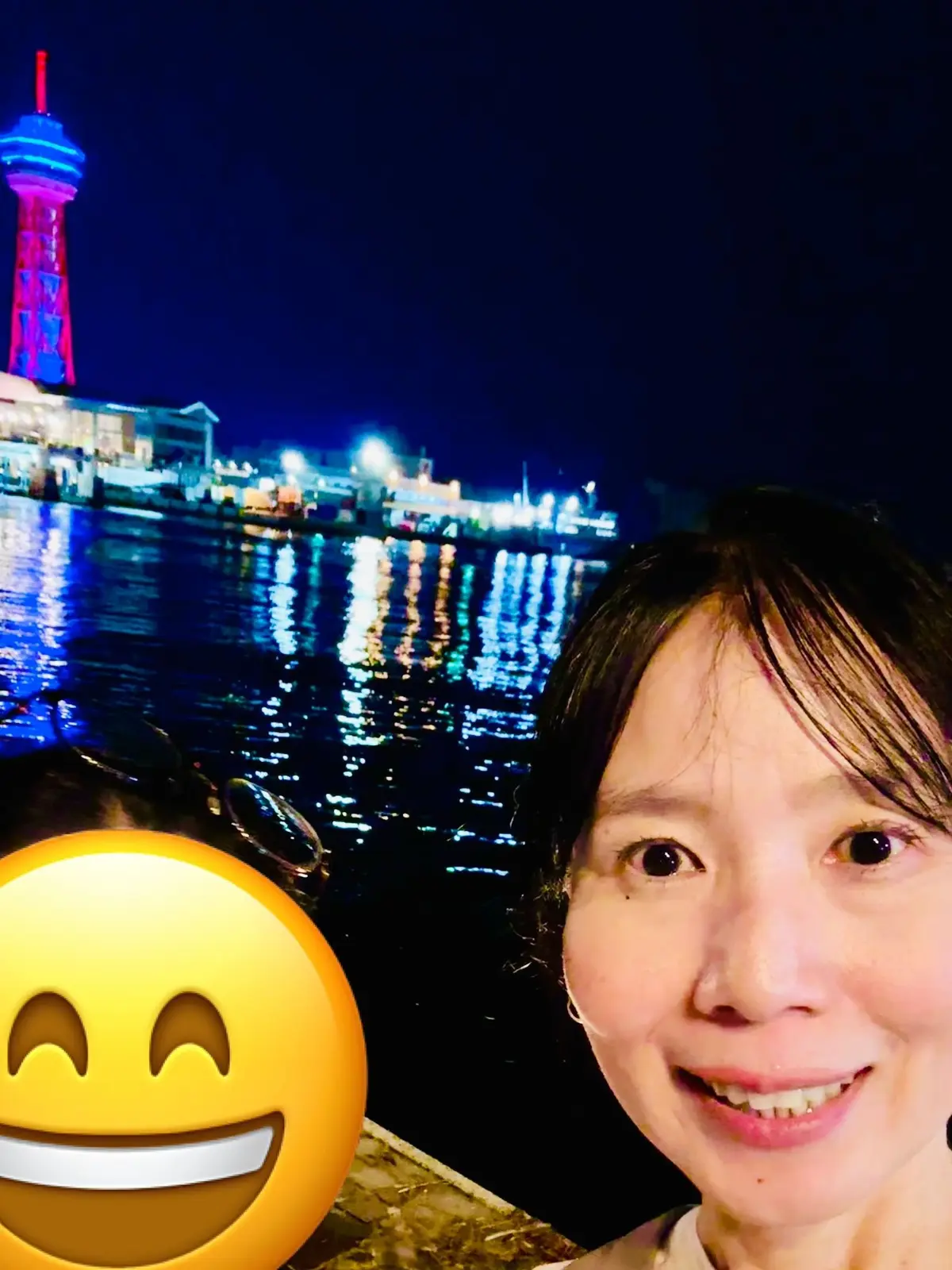 夜景をバックに笑顔の女性。手前に笑う顔の絵文字、背景にはライトアップされた観覧車と遊園地。水面に光が反射している。