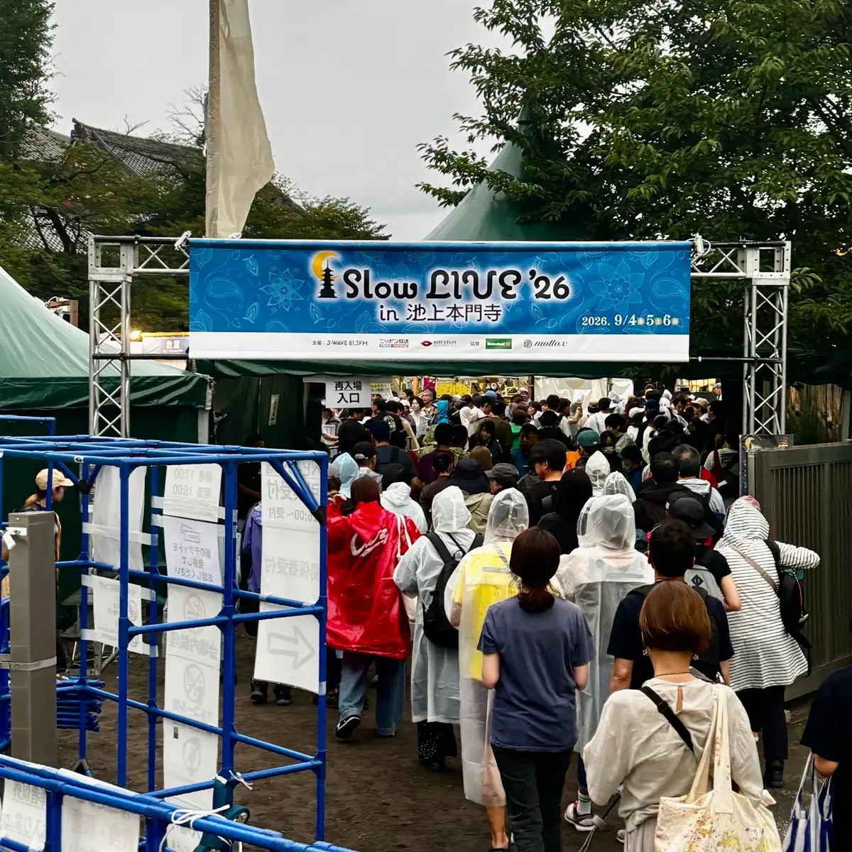 屋外イベント会場の入り口付近。多くの人がゲートを通ろうと並んでおり、青い柵で区切られている。背景には緑豊かな木々とイベントの看板が見える。