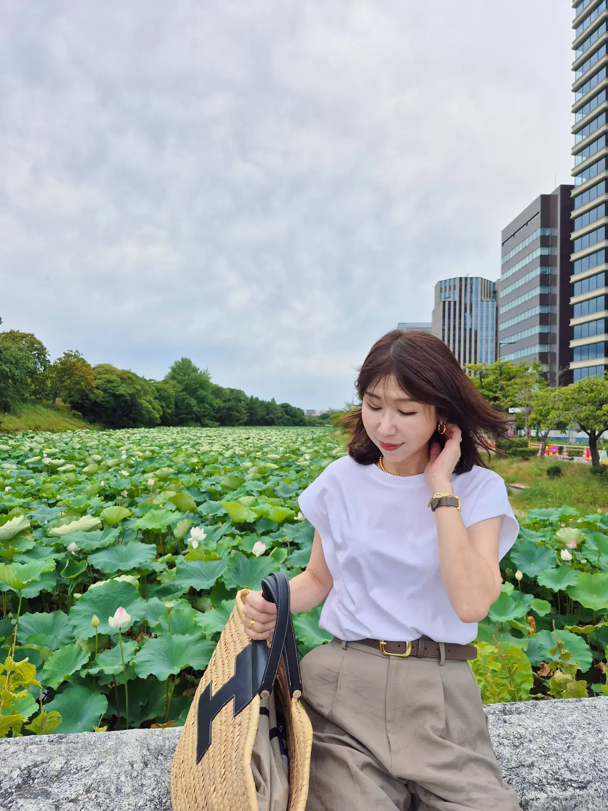 水辺に咲く睡蓮の池を背景に、白いTシャツとベージュのパンツを着た女性が座っている。右手にバッグを持ち、髪をかき上げている。背後には高層ビルが見える。