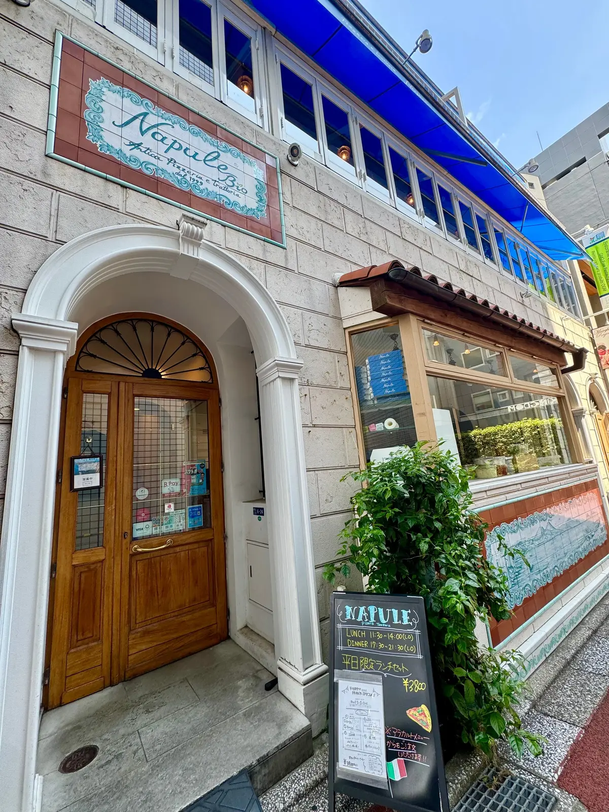イタリア料理店「ナプリーズ」の入り口。石造りの建物で、アーチ型の扉と窓、装飾的な装飾が施されている。看板にはランチとディナーの時間、お得なセットメニューが記載されている。