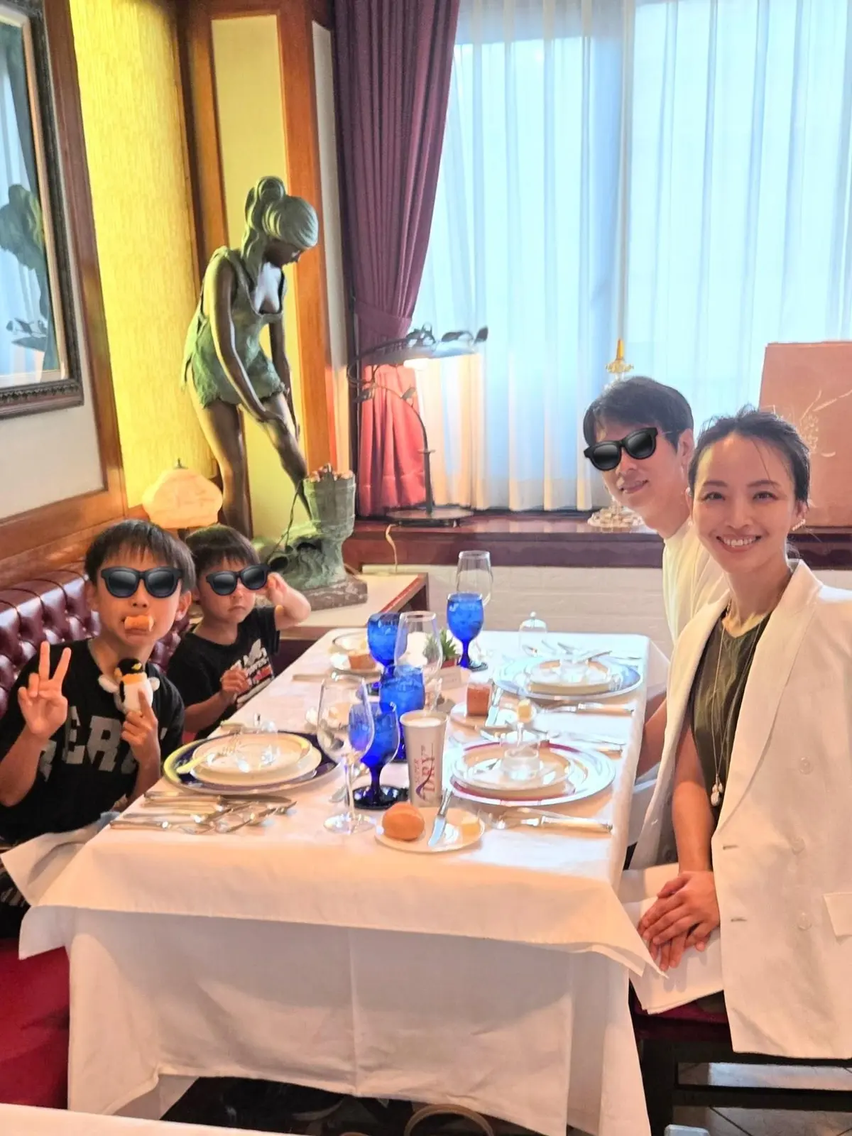 白テーブルを囲み、笑顔の家族4人が写っている。左端にはサングラスをかけた少年がピースサイン、右隣は父親、右端は母親。背景には彫刻とカーテン、窓から光が差し込んでいる。