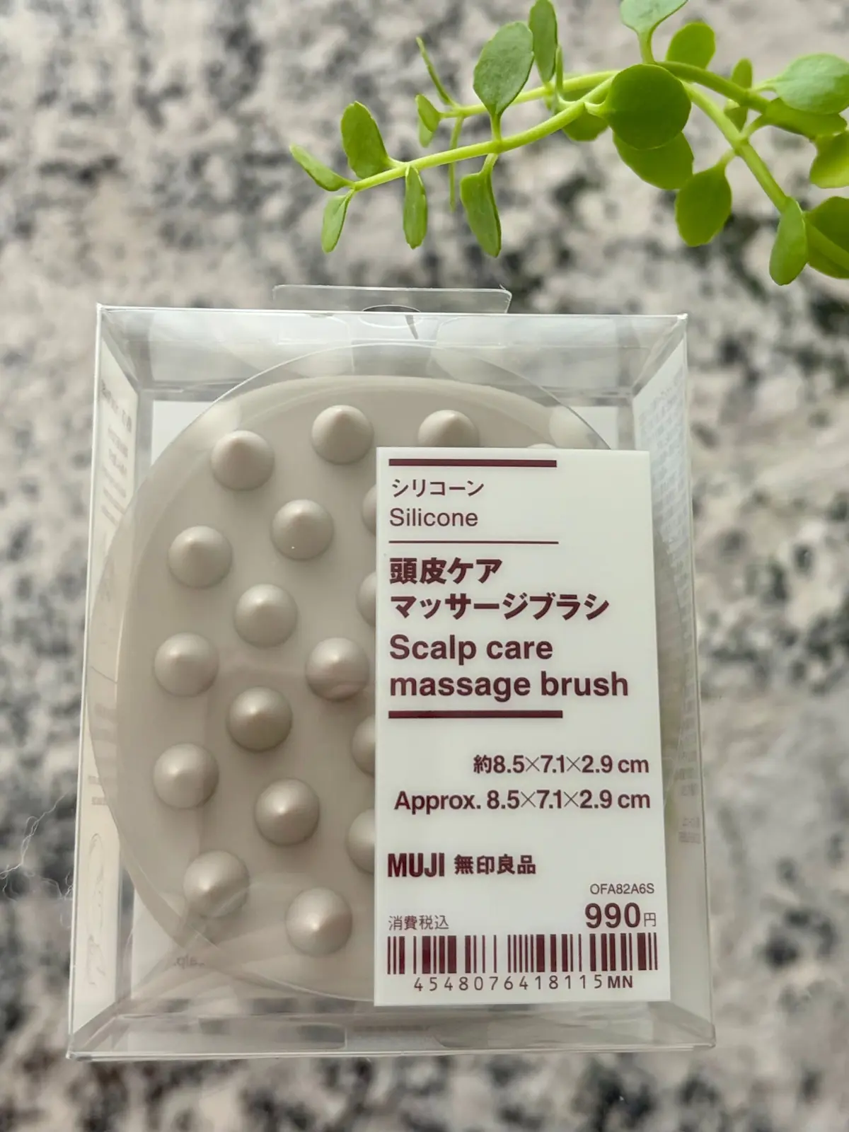 透明なケースに入った、淡いピンク色のシリコン製頭皮マッサージブラシ14個。MUJIの製品で、サイズは約8.5x7.1x2.9cm。背景には緑の植物が写っている。