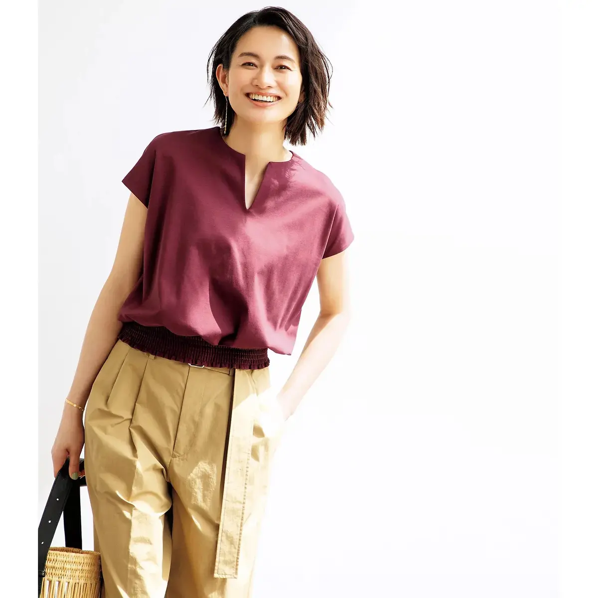 女性がマスタード色のワイドパンツと深紫のVネックカットソーを着て、左手で編みかごバッグを持ち、右手は腰に置いた姿勢で、明るい背景に立っています。