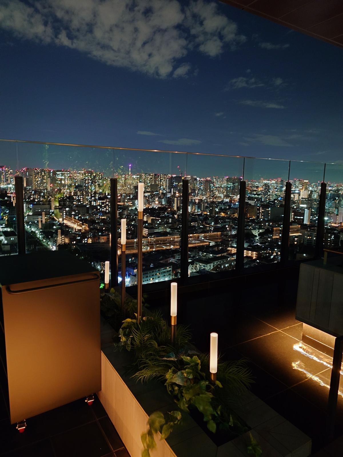 夜景を望む The TRAVELERS HOUSE ROOFTOP BAR のバルコニー。ガラス張りの手すみが視界を遮る。手前に植栽があり、ライトアップされている。遠景には都市の光が広がり、雲が浮かぶ夜空。