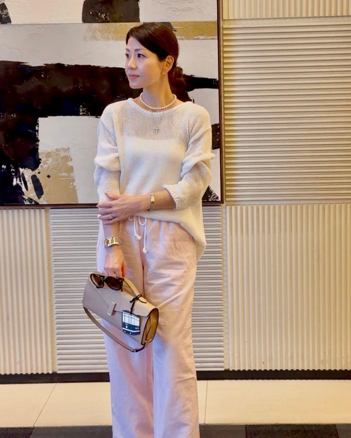 女性が白いニットセーターとベージュのワンピースを着て、抽象画の前で立ち、左手でサングラス付きのトートバッグを持っている。