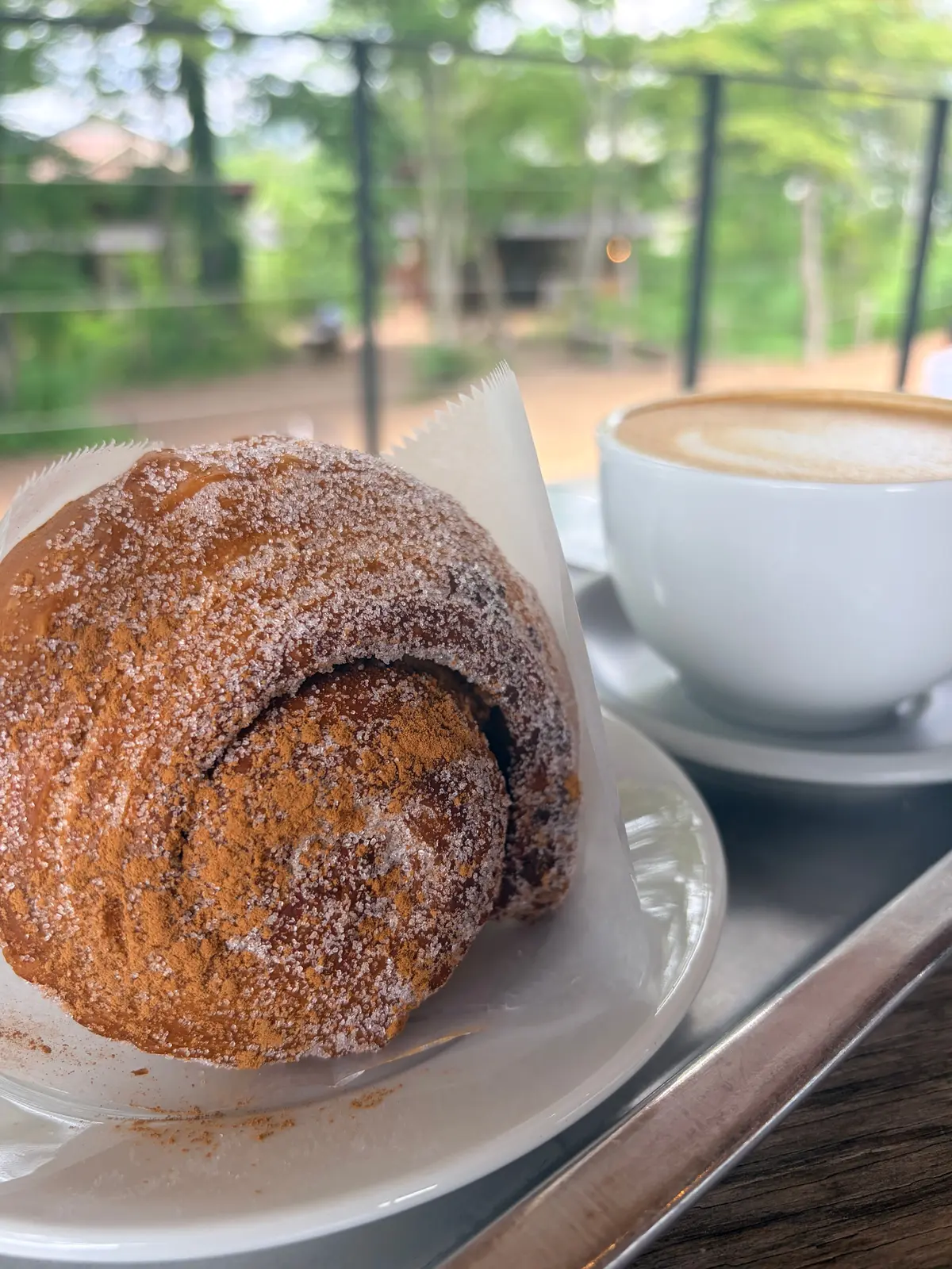 白い皿に砂糖をかけたクロワッサンと、白いカップに入ったカフェラテ。窓の外には緑豊かな風景が見える。