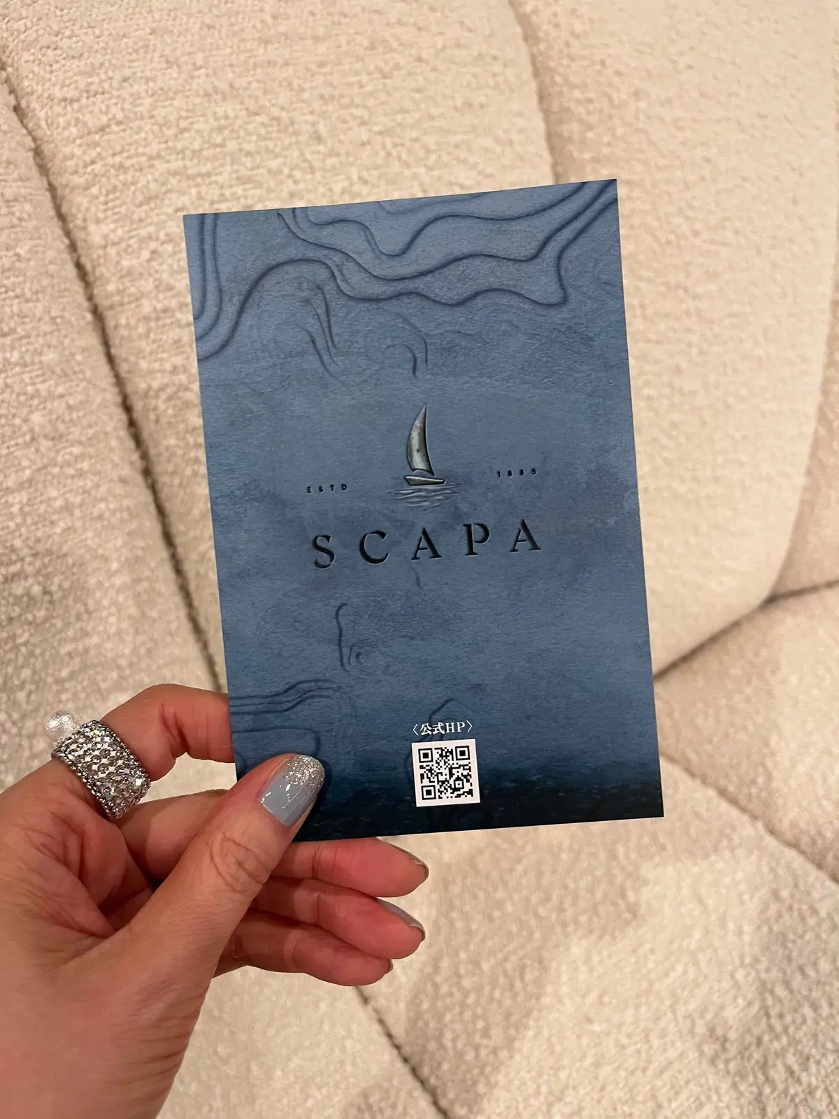 青みがかったグレーの背景に、波の模様とヨットのイラストが描かれたカード。中央に「SCAPA」のロゴと、ESTD 1889の文字が配置されている。右下にはQRコードと「公式HP」の文字がある。