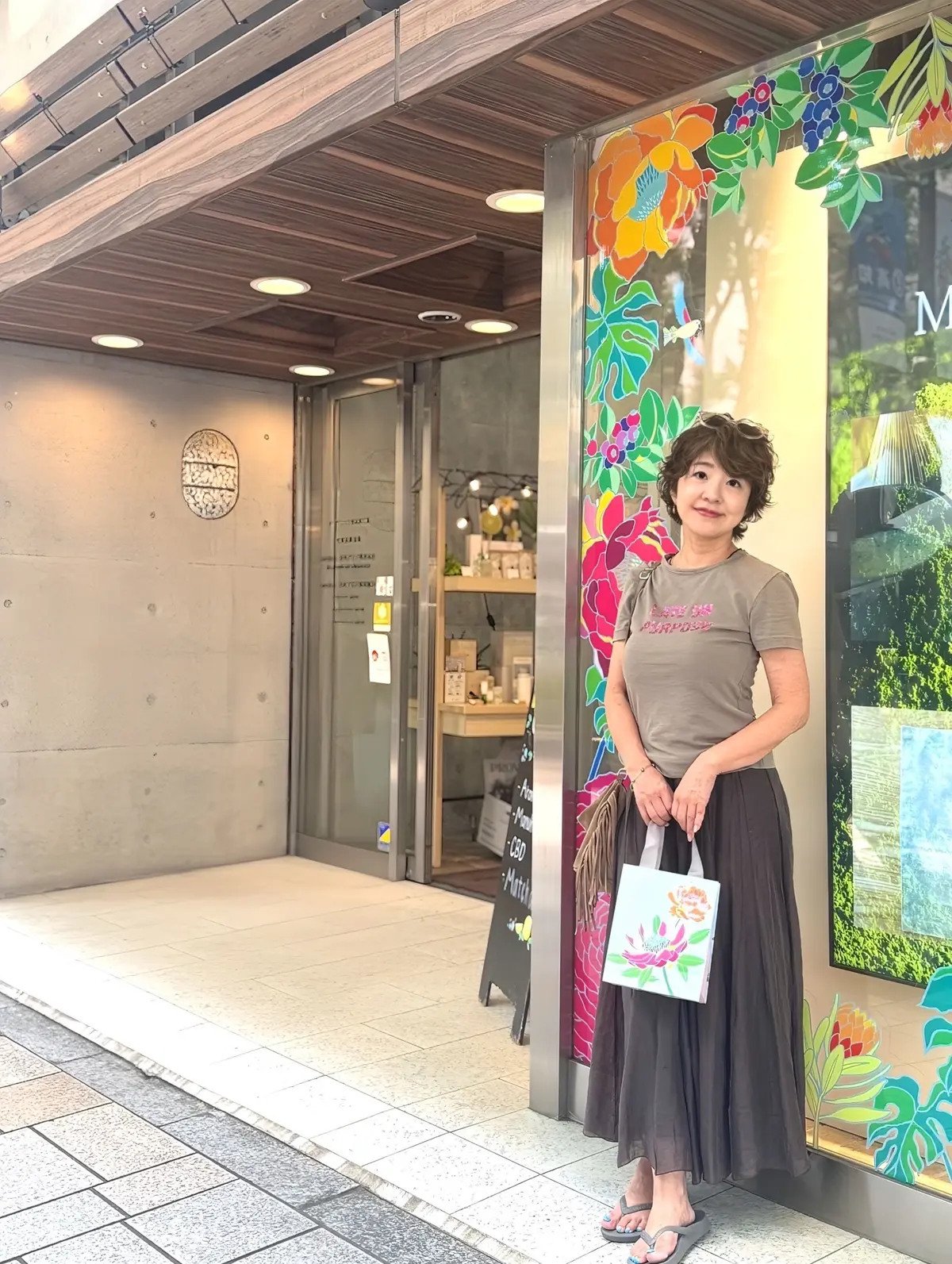 茶色のTシャツとグレーのロングスカートを着た女性が、鮮やかな花柄の壁の前に立っている。背景には、木製のドアと窓のあるお店が見える。女性は右手にトートバッグを持ち、リラックスした表情をしている。
