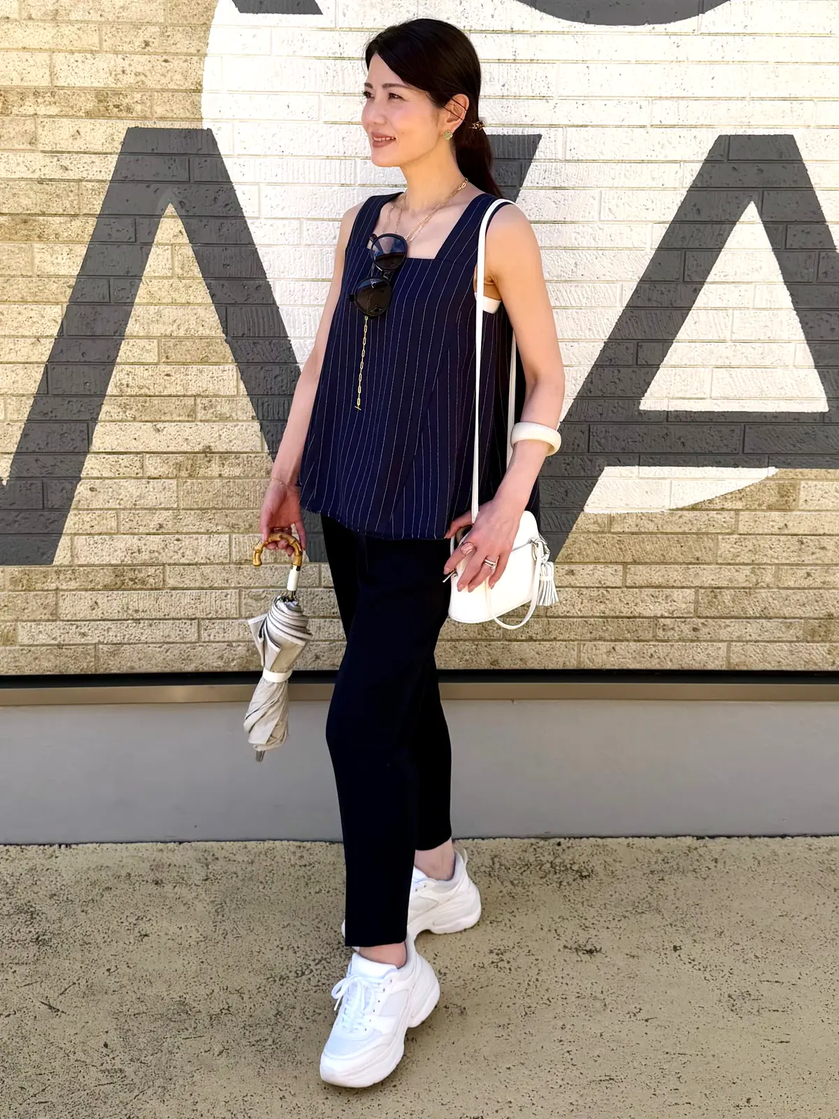 青いピンストライプのトップスに黒のパンツ、白いスニーカーを着用した女性が立っている。右手に白い傘を持ち、左腕には白いショルダーバッグをかけている。背景にはグレーと白の幾何学模様の壁がある。