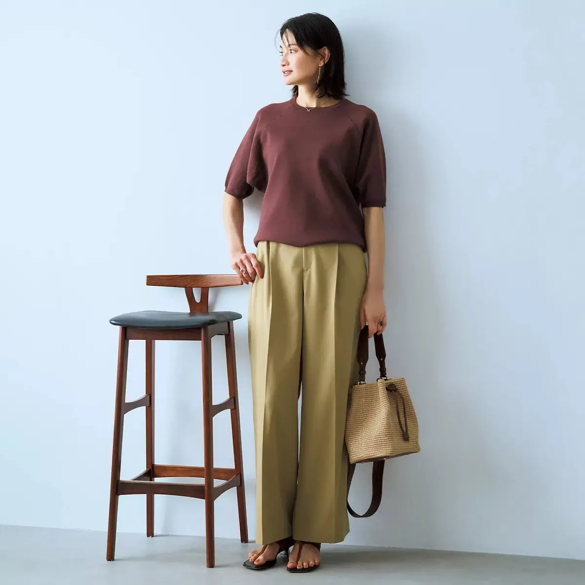 ブラウンのニットトップスに、ベージュのワイドパンツを合わせた女性。右手にストロー製のバッグを持ち、木製のスツールを背に立っている。