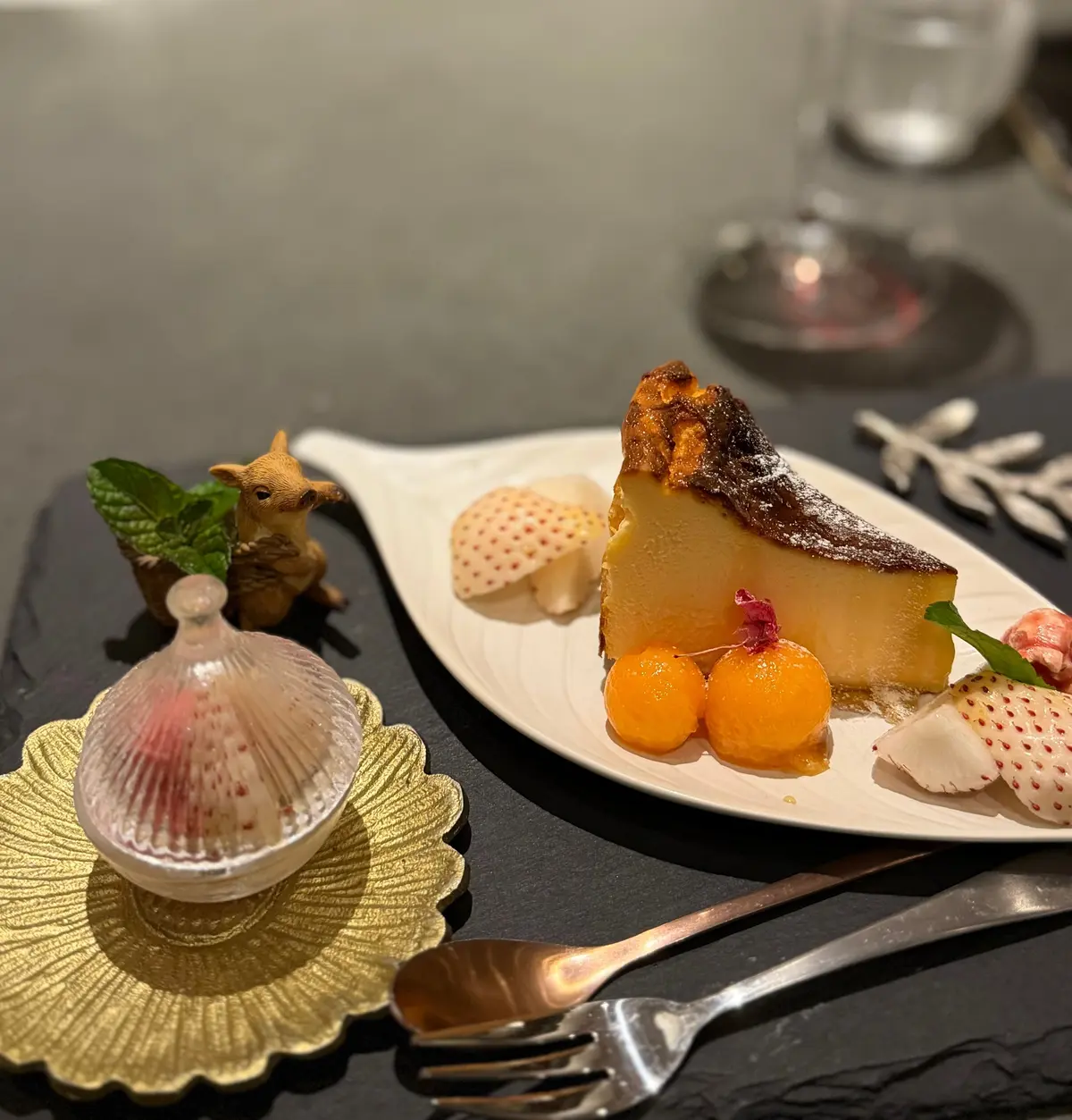 黒いテーブルに置かれた白い皿に、バスクチーズケーキの切れ端とフルーツ、飾り付けが施されている。金色のコースターに乗ったガラスの器と、小さなリスの置物も写っている。