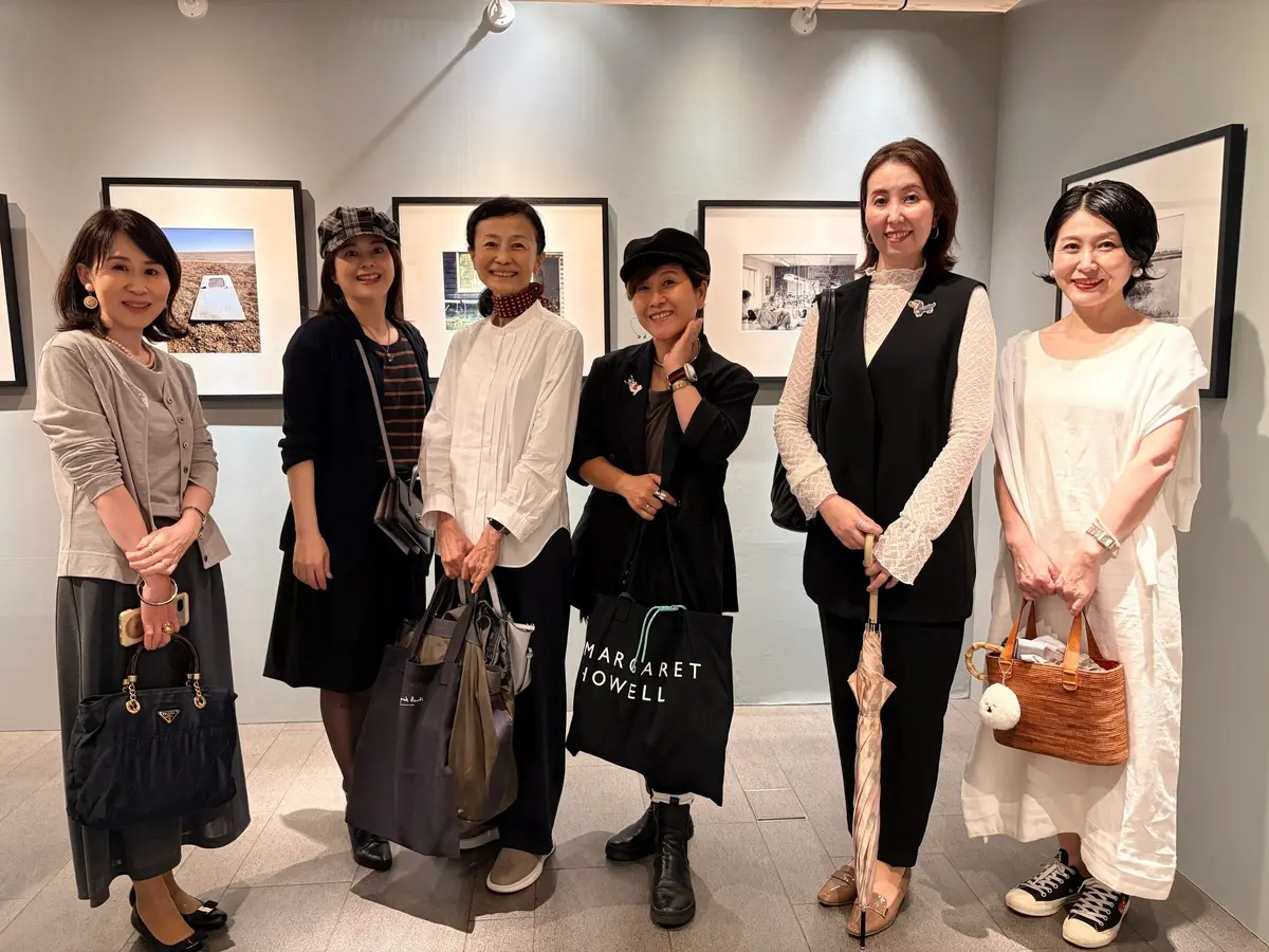 ギャラリーで写真作品を鑑賞する6人の女性。白い壁の前に立ち、笑顔で会話を楽しんでいる。左から、グレーのニットを着用した女性、黒い服の女性、赤い襟のシャツを着た女性、黒い帽子をかぶった女性、ベストを着た女性、白いシャツを着た女性。
