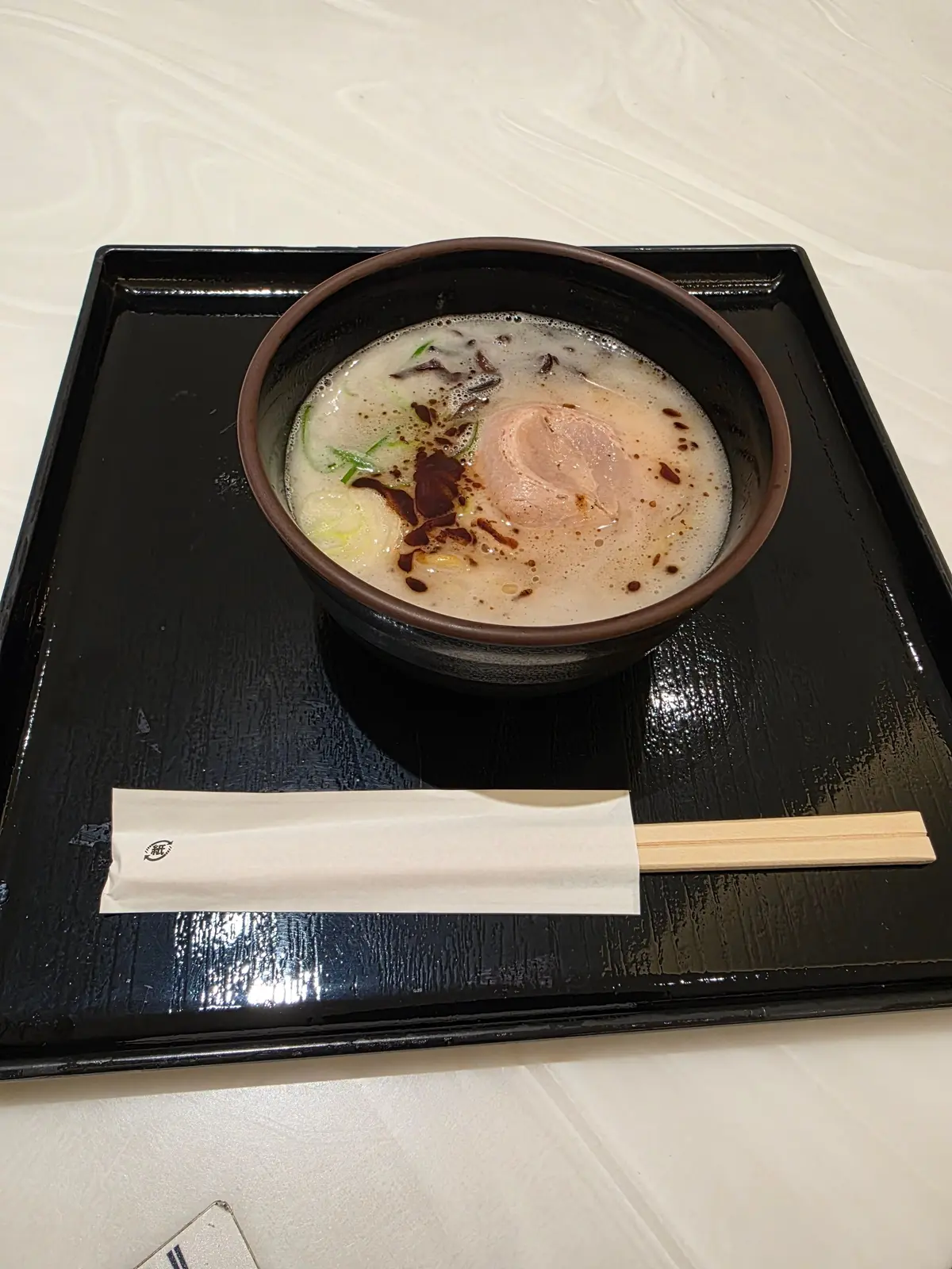 黒い漆塗りの木製プレートに盛り付けられたラーメン。茶色の器に白いスープ、麺、薄切り肉、ネギ、醤油ダレが浮かんでいる。箸と包み紙が添えられている。
