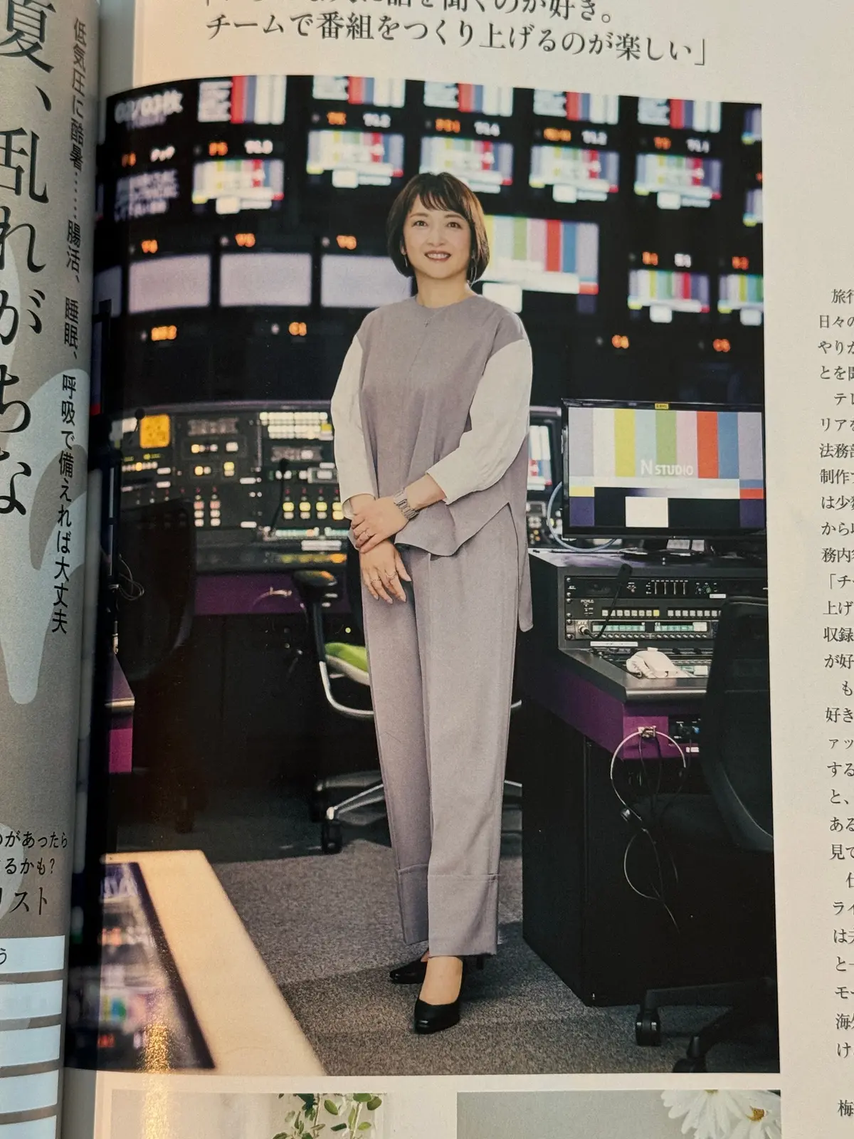 女性がテレビスタジオで立っており、複数のモニターと制御パネルが背景に並んでいる。彼女はグレーのスーツを着て、両手を腰に置いている。