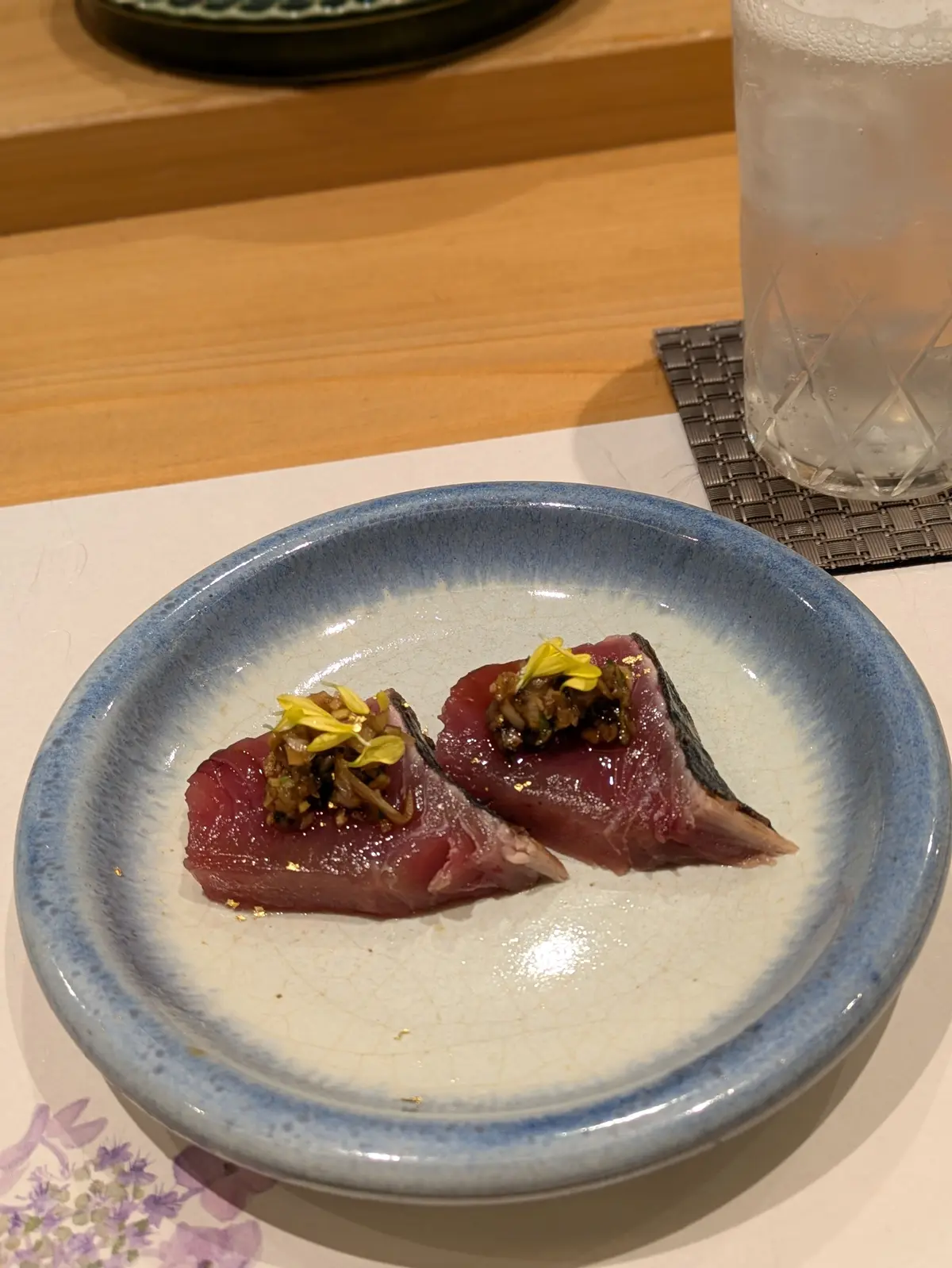 青い縁の白い陶器の皿に、2つの赤身魚の切り身が並び、黄色い花と細かく刻んだ野菜のトッピングがのっている
