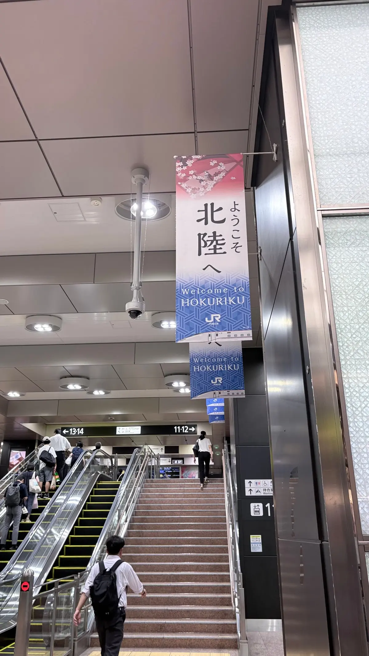 駅構内の階段とエスカレーター。上部には「Welcome to HOKURIKU」と書かれた看板が掲示され、ピンクと青の配色が特徴的。手すりを背に人々が階段を上っている。