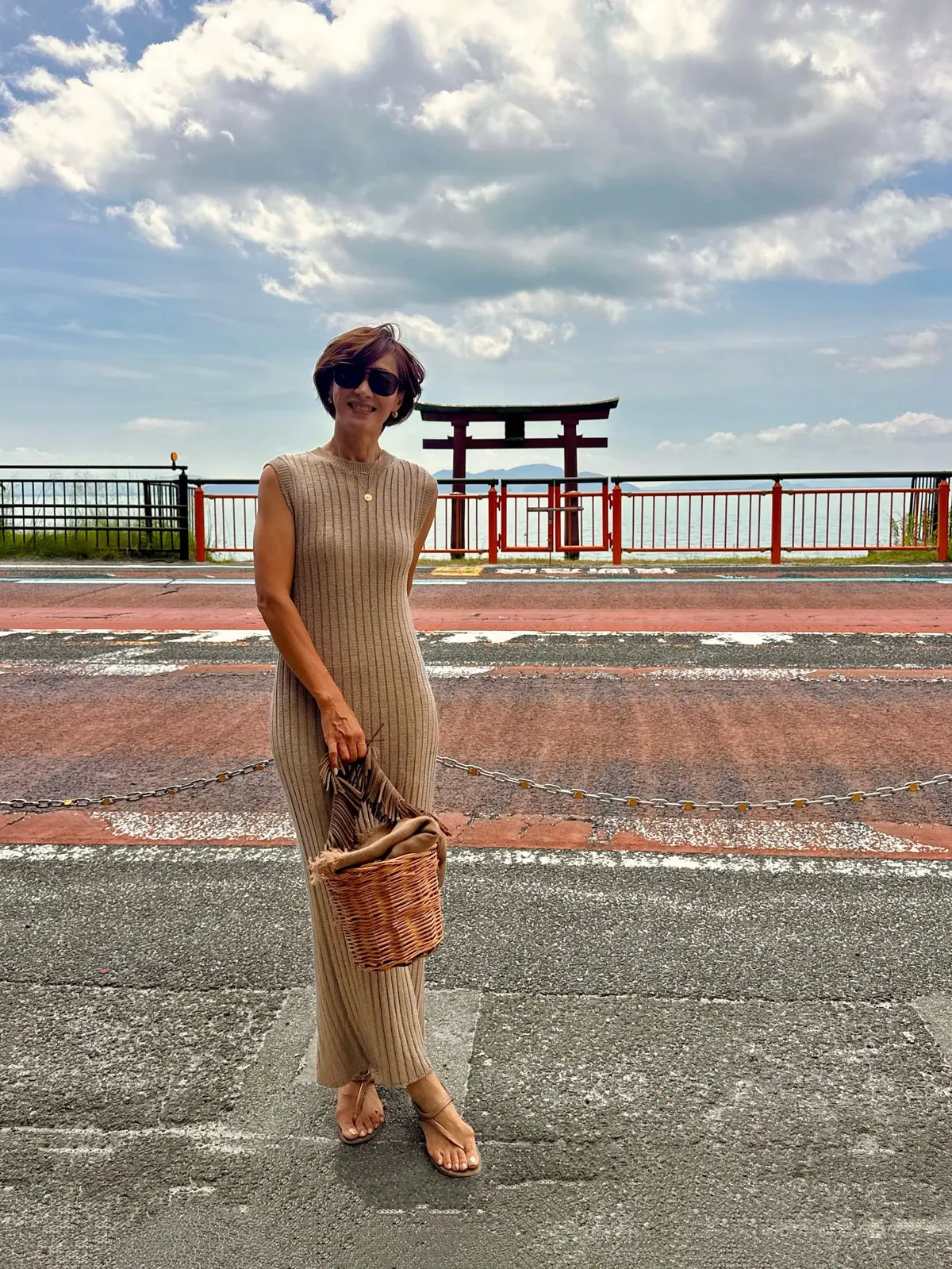ベージュのニットワンピースを着た女性が、海を望む赤い鳥居の前でポーズをとっている。右手にカゴバッグを持ち、サンダルを履いている。背景は曇り空。