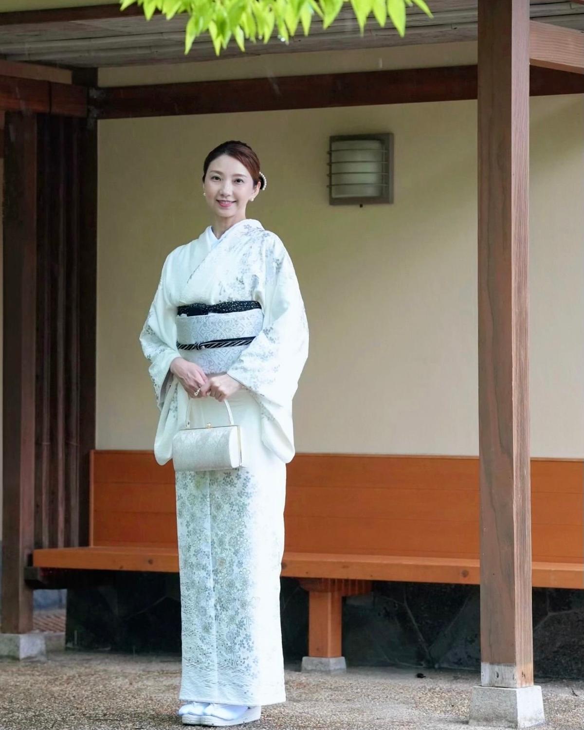 白い和服を着た女性が、木造の建物の軒下に立ち、微笑んでいる。手には小さな手提げ袋を持っている。