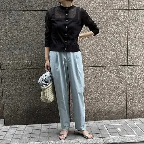 黒い透け感のある長袖トップスに、淡いブルーのプリーツパンツを合わせた女性。シルバーのサンダルを履き、麦わら風のバッグを持つ。背景はグレーのタイル壁。
