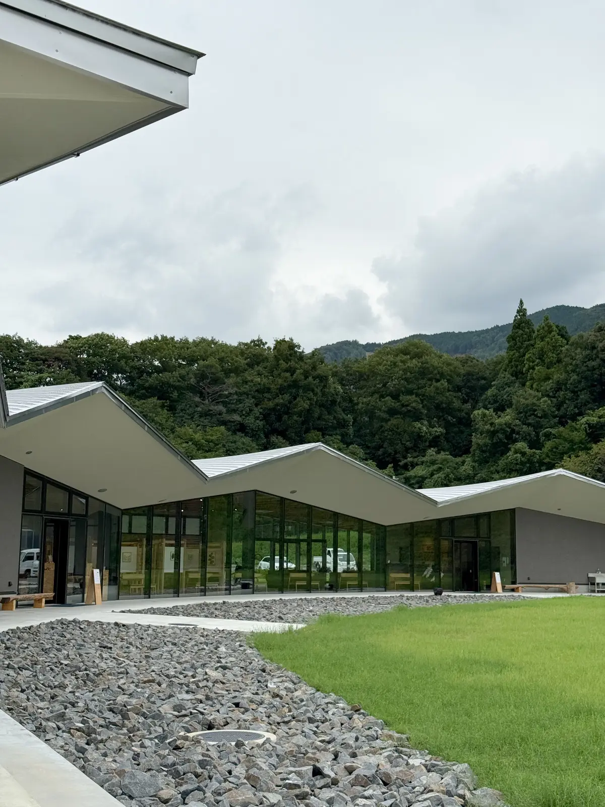緑豊かな丘に面した、グレーと白のモダンな建築物。緩やかな勾配の屋根が連続し、大きな窓が並ぶ。手前には小石敷きの通路と芝生が広がり、自然と調和した景観。