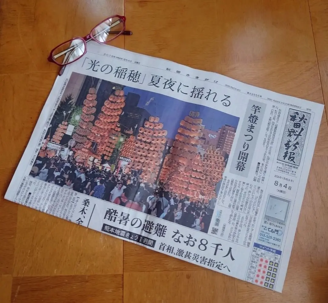 木製のテーブルに置かれた日本の新聞。「光の稲穂」と大きく書かれた見出しの下に、夜空に浮かぶ多数の提灯の写真が掲載されている。右上に「読売新聞」のロゴ、下部には電話番号や広告が表示されている。